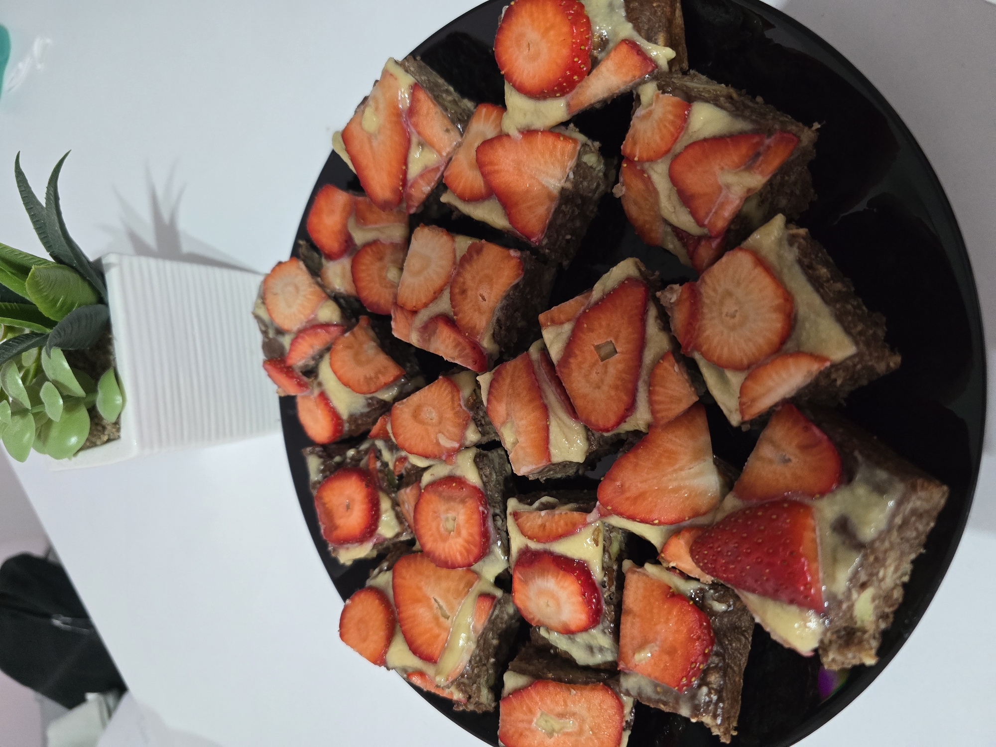 The image shows a black plate filled with square-cut pastries topped with slices of strawberries and a light-colored cream. A small, square white pot containing a green artificial plant is situated near the plate on a white surface. There are no people visible. The location is unspecified but likely indoor given the controlled light and presence of an artificial plant. There is no indication that it is in Burgas, Bulgaria.