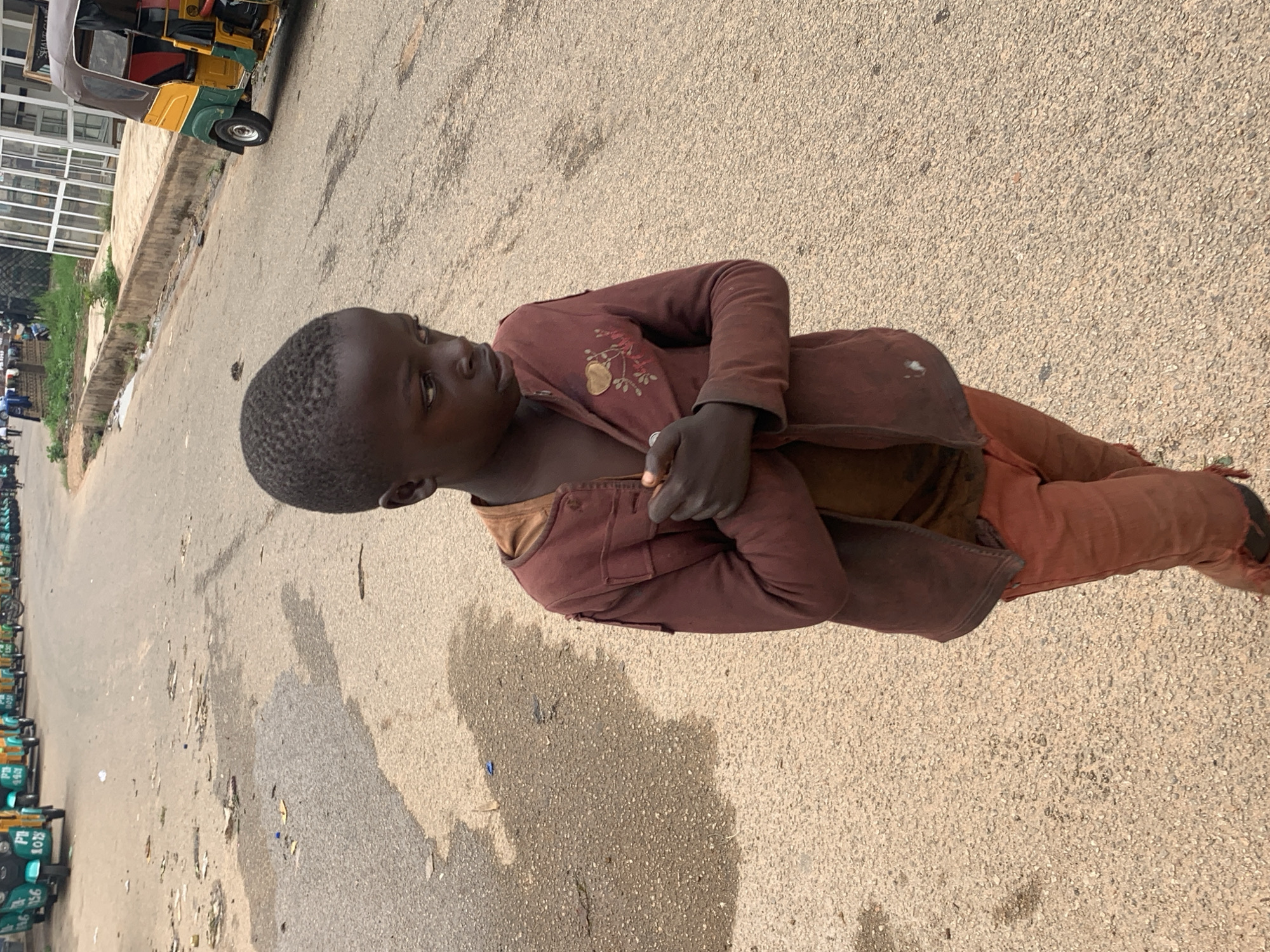 An African boy stands on a city street in Jos, Nigeria. He wears an unbuttoned maroon jacket over a t-shirt and maroon pants. His arms are folded across his chest. The street is paved and shows wear and tear. Vehicles are parked in the distance.