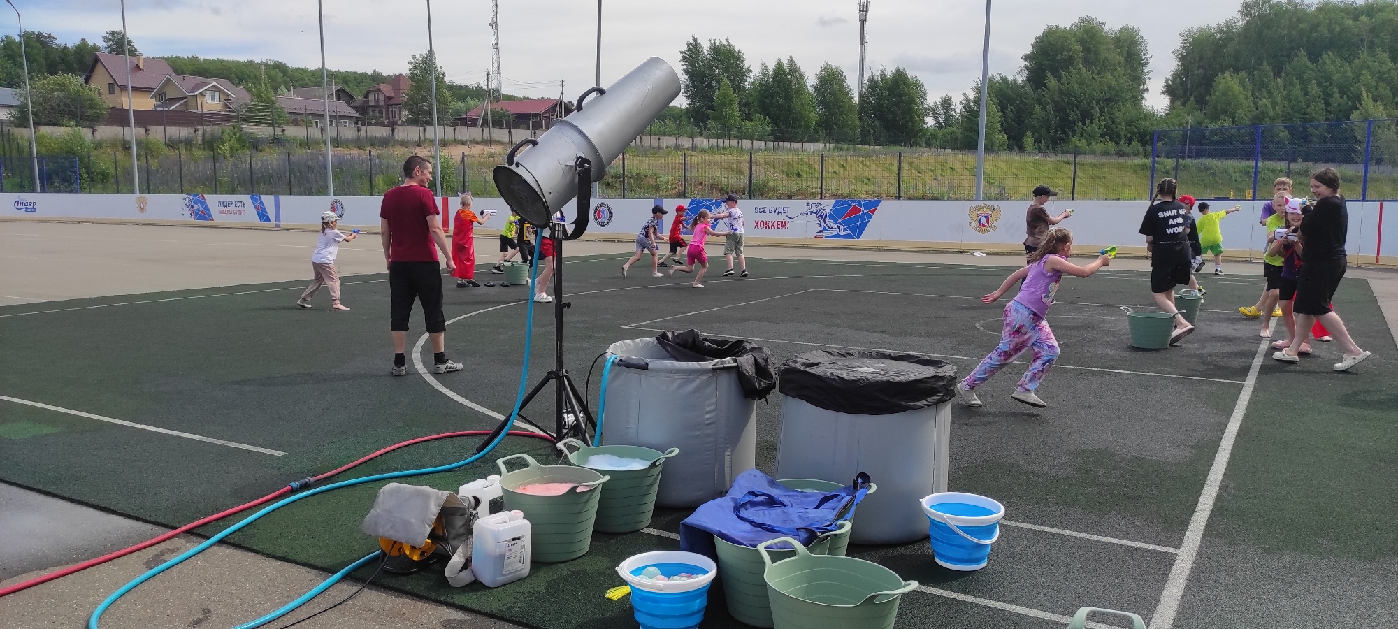 In Noviy, Russia, a group of children is engaged in a water gun fight on an outdoor artificial turf sports field. The scene unfolds under a cloudy sky, with a line of trees and houses visible in the background, suggesting a suburban or park-like setting. In the foreground, several large grey and green buckets are filled with water, some containing colored liquid. A large, metallic, cannon-like device on a tripod is positioned to the left of the frame, connected by blue and red hoses to what appears to be a water source. Several children, dressed in casual summer clothing, are actively participating in the water fight. Some are aiming water guns, while others are running and dodging. A few adults are also present, observing the activity. The sports field is marked with white lines. The overall atmosphere is one of energetic, recreational play.