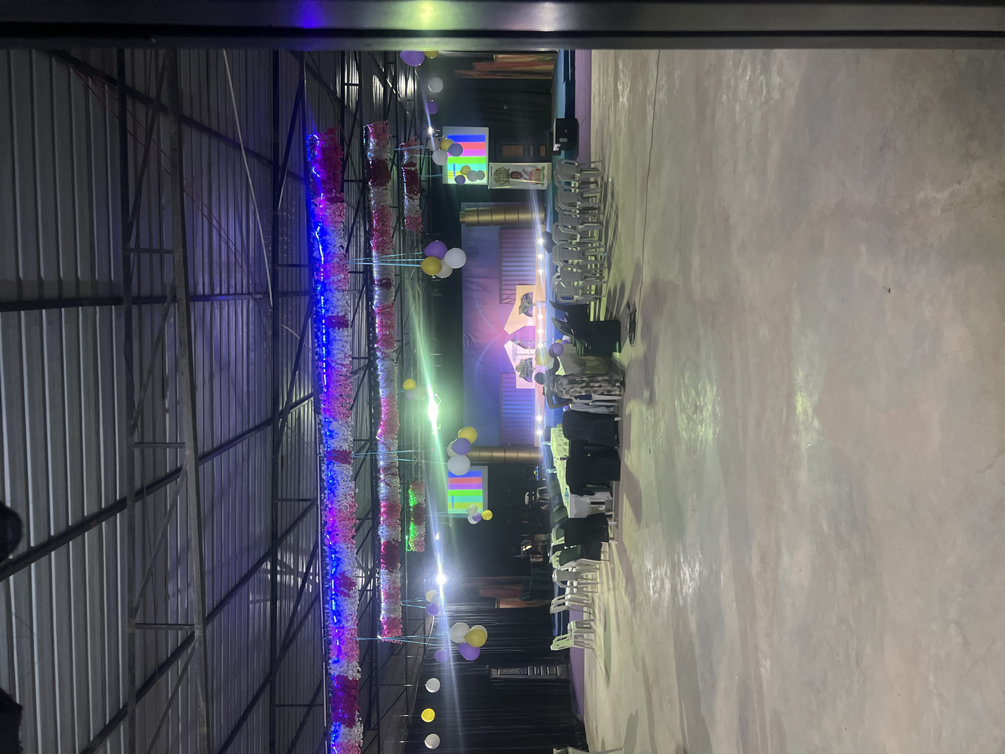 The image depicts an indoor event space in Durumi, Nigeria. The scene appears to be set up for a gathering, possibly a ceremony or a conference. The main area features a stage at the back with a large screen displaying a colorful test pattern, suggesting it's a video display. There are seating arrangements in the foreground, with rows of chairs and tables covered in white and black cloths. Decorations, including strings of colorful flowers and balloons, hang from the ceiling, which is constructed with exposed metal beams. Several people are visible, some seated at tables and others standing. The lighting suggests an evening or indoor event. The floor is concrete. The overall impression is that of a functional, decorated event venue prepared for attendees.