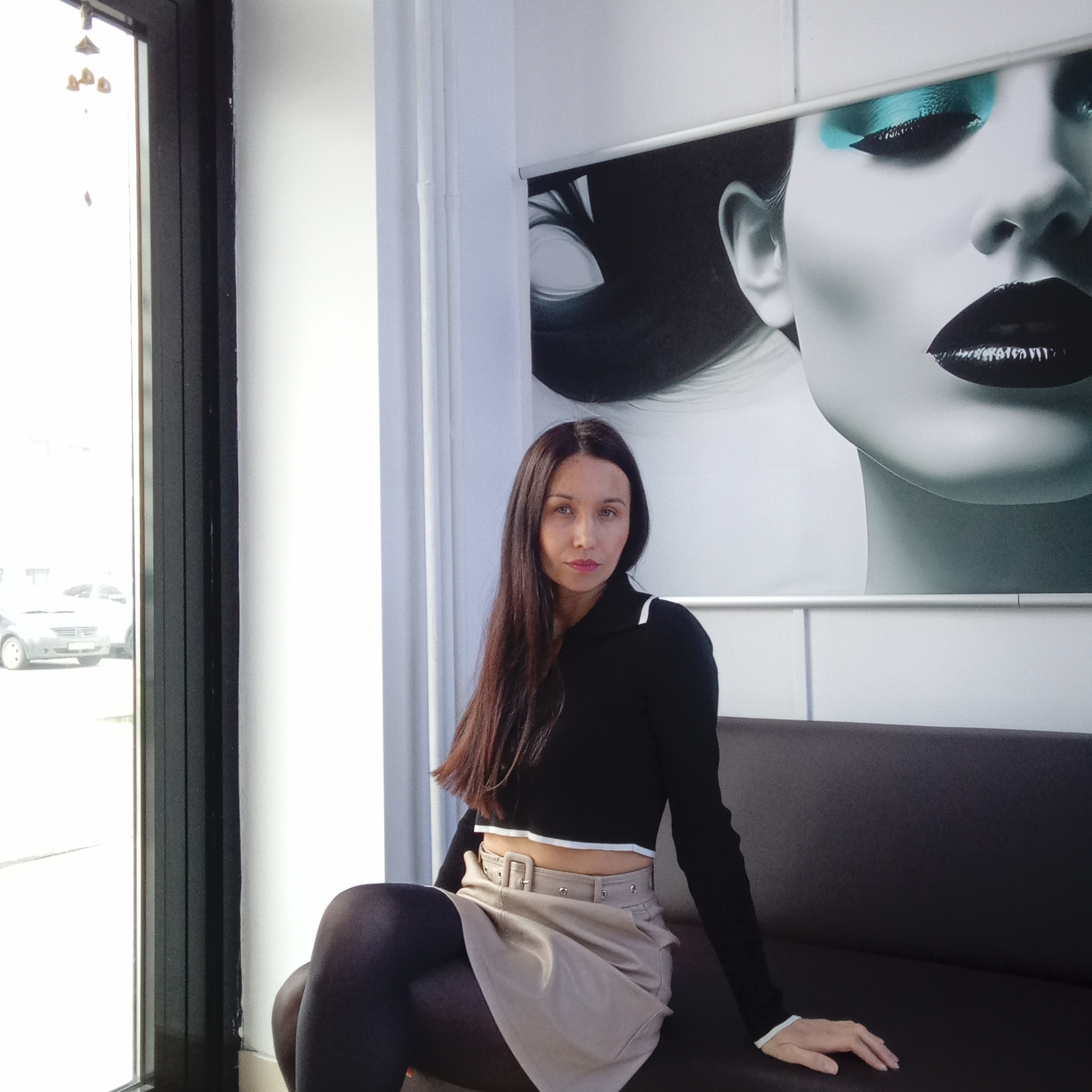 A woman with long, straight dark hair is seated on a dark brown, leather-like couch in an indoor setting, in Izhevsk, Russia. She is wearing a black long-sleeved crop top with a white collar and white trim on the cuffs, a light beige mini skirt with a buckled belt and metallic studs, and opaque dark tights. Her face is turned slightly towards the viewer, displaying a calm, neutral expression, with subtle light pink lipstick.
Behind her, a large, prominent poster on the wall depicts a stylized close-up of a woman's face, characterized by bold black lips and vibrant aqua eyeshadow. The poster is framed by a thin silver bar at the top. To the left, a large window allows bright daylight to illuminate the room, offering a view of a street outside with parked cars. Small, golden-colored, bell-like ornaments are visible hanging near the top of the window frame. The strong natural light indicates it is daytime. The overall scene suggests a modern, clean interior, possibly a waiting area, office, or salon. No discernible text is visible within the image.