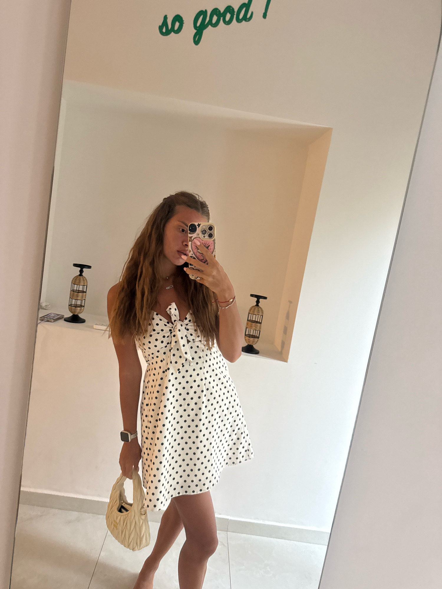 In Stavros, Greece, a young woman with long, wavy brown hair takes a mirror selfie. She is wearing a white dress with black polka dots and a bow at the neckline. She is holding a small, cream-colored purse in her left hand. On her left wrist, she wears a silver watch. Her right hand is holding a smartphone, which is reflecting the image. In the background, on a shelf, are two decorative wicker lanterns. Above the mirror, the words "so good!" are written in green cursive. The wall behind her is white, and a portion of a light-colored floor is visible.