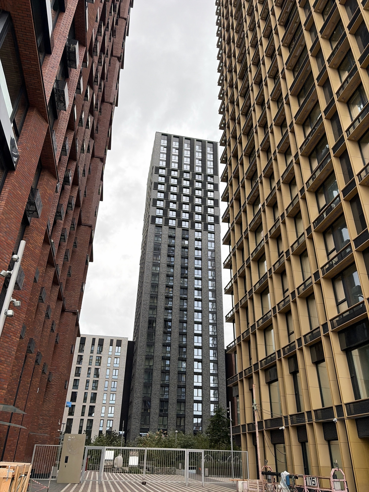 The image depicts an exterior view of modern high-rise buildings in an urban setting. In the foreground, two distinct buildings flank a central, taller structure. The building on the left is clad in dark red brick with a textured facade. To the right, a building with a tan or golden facade features a grid-like pattern of windows and vertical supports. The central building is a very tall, slender skyscraper with a gray facade and numerous rectangular windows arranged in a uniform pattern. In the background, another, shorter white building with a similar window pattern is visible between the red brick building and the central skyscraper. The scene is set under an overcast sky. A white metal fence runs across the lower portion of the image, with what appear to be construction materials and equipment to the left and right. This scene is located in Moscow, Russia.