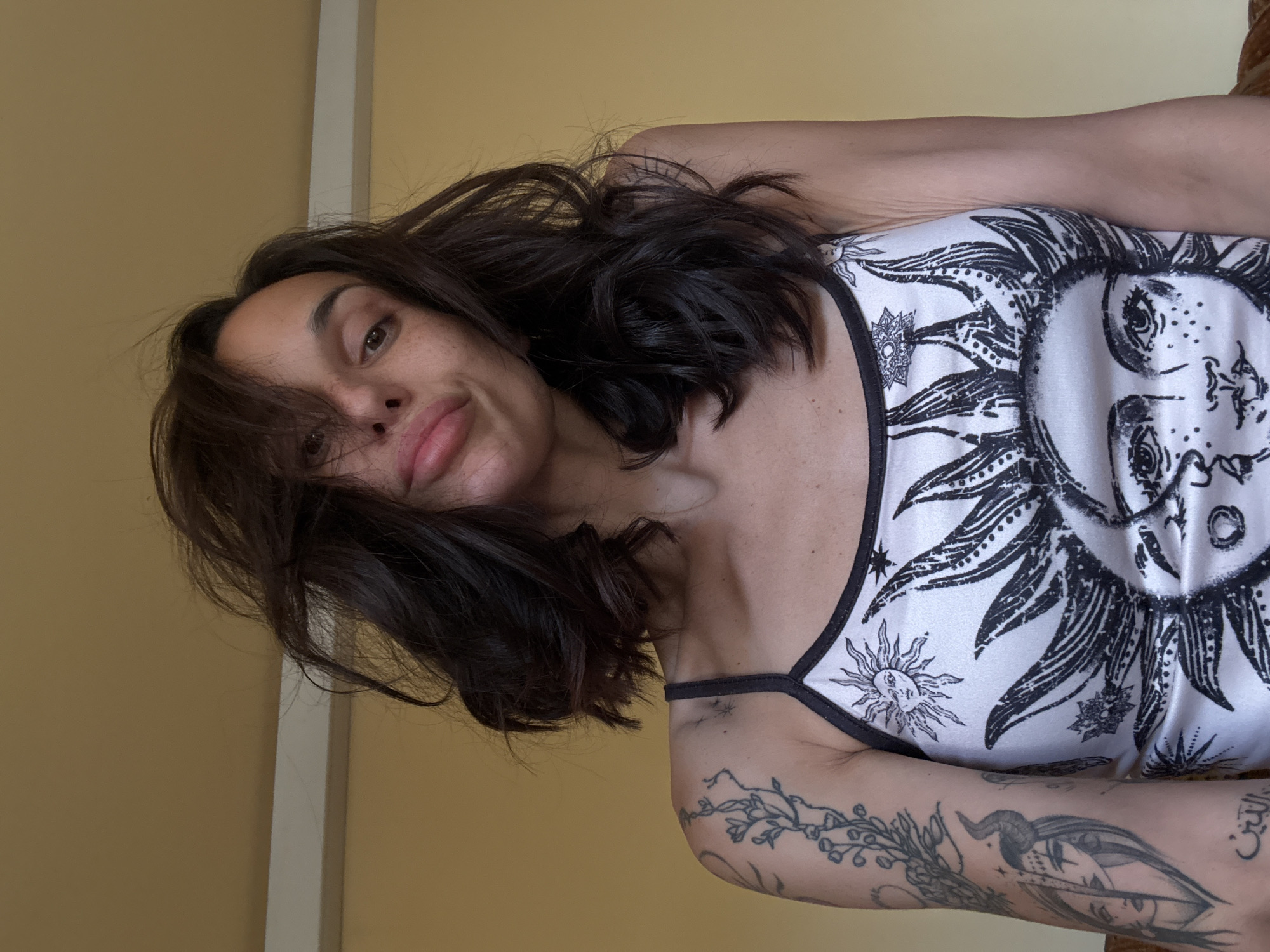 A woman with dark brown hair and freckles is pictured indoors. She is wearing a white tank top with a black and white sun design. The woman has tattoos on her left arm. The wall behind her is a light yellow color. The location of the image is unknown.