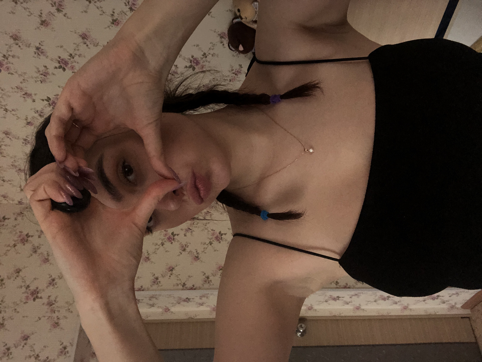 The image displays a female individual situated indoors in Noviy, Russia. The subject is positioned centrally, facing forward, with her hands raised to form a heart shape in front of her face, partially obscuring her eyes. Her lips are pursed into a pouting expression. She possesses dark hair styled into two braids, with a blue elastic band securing the braid on the left side and a purple elastic band on the right. Her fingernails are long and painted with a light, reflective polish. She is attired in a black spaghetti-strap top and wears a thin, gold-toned necklace featuring a small, clear gemstone pendant. The background consists of a wall covered in light-toned wallpaper adorned with a pattern of pink and purple floral clusters. To the left, a segment of a light-colored wooden door frame and a door with a metallic doorknob are visible. To the right, two plush toys, one brown and one lighter in color, are situated on a surface behind her right shoulder. The scene is illuminated by artificial indoor lighting.