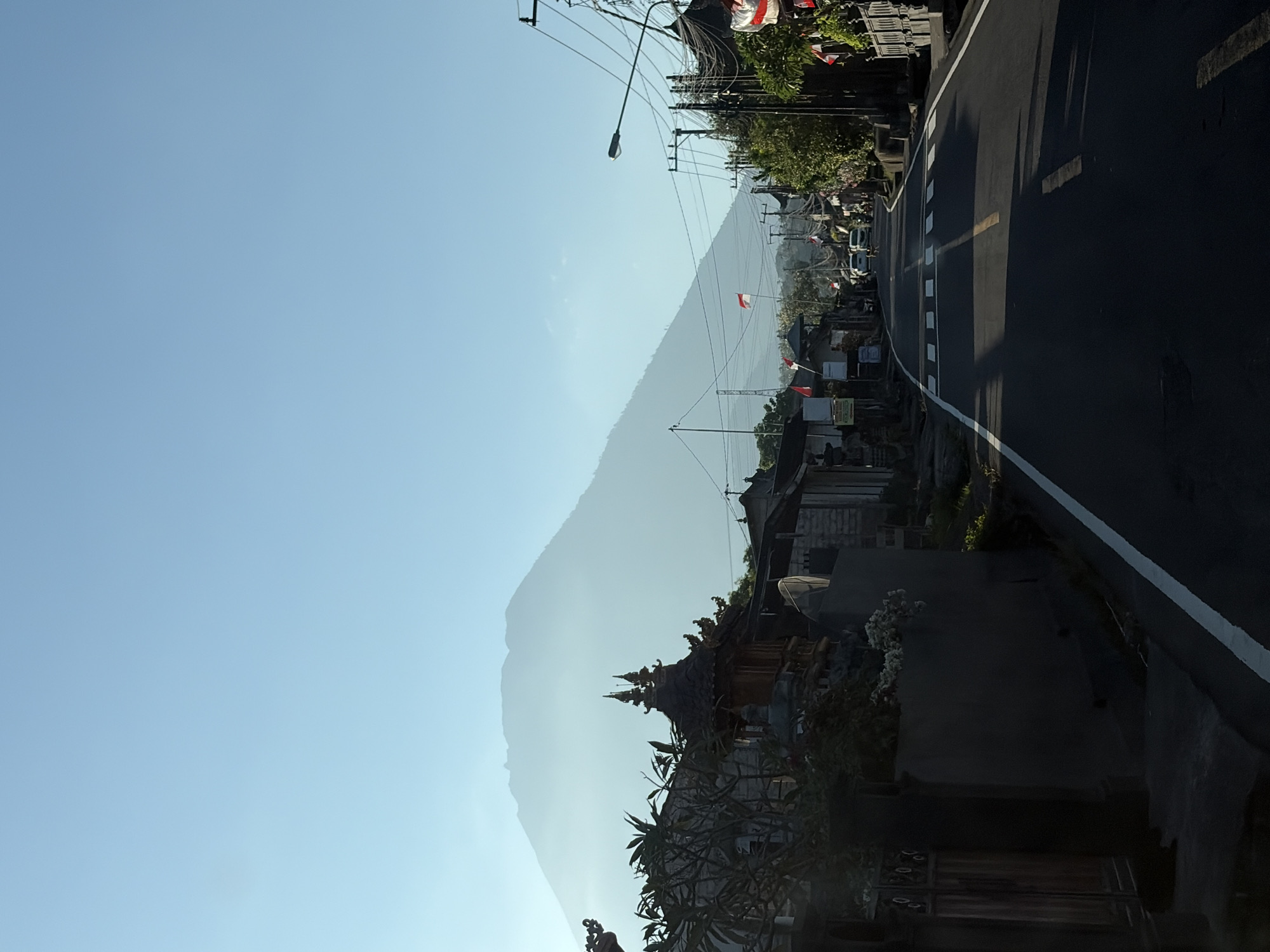 The image depicts a road in Karangasem, Indonesia, with a prominent, misty mountain in the background. The road is paved and asphalted, with a white line marking the edge and a yellow line dividing the lanes. There are several small buildings lining the street, characterized by traditional Balinese architecture with intricate roofs and stone walls. Small Indonesian flags are visible, some hanging from poles and others attached to buildings, suggesting a local celebration or national holiday. A few cars are parked or driving on the road. The sky is clear and bright blue, with the sun casting shadows on the road. The overall atmosphere is serene, with a sense of rural tranquility enhanced by the presence of the majestic mountain.