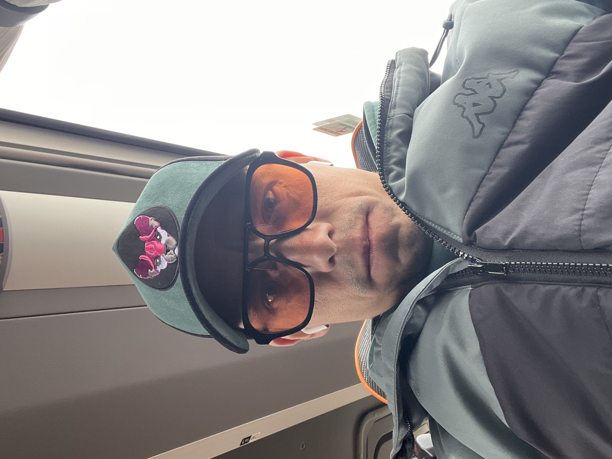 A man wearing sunglasses, a green cap with a floral emblem, and a Kappa jacket is captured in an upward-angled photograph. He appears to be on a train or bus due to the presence of a window and interior paneling in the background. The location is indeterminate due to the lack of specific identifiers relating to the city of Chaikovsky, Russia.