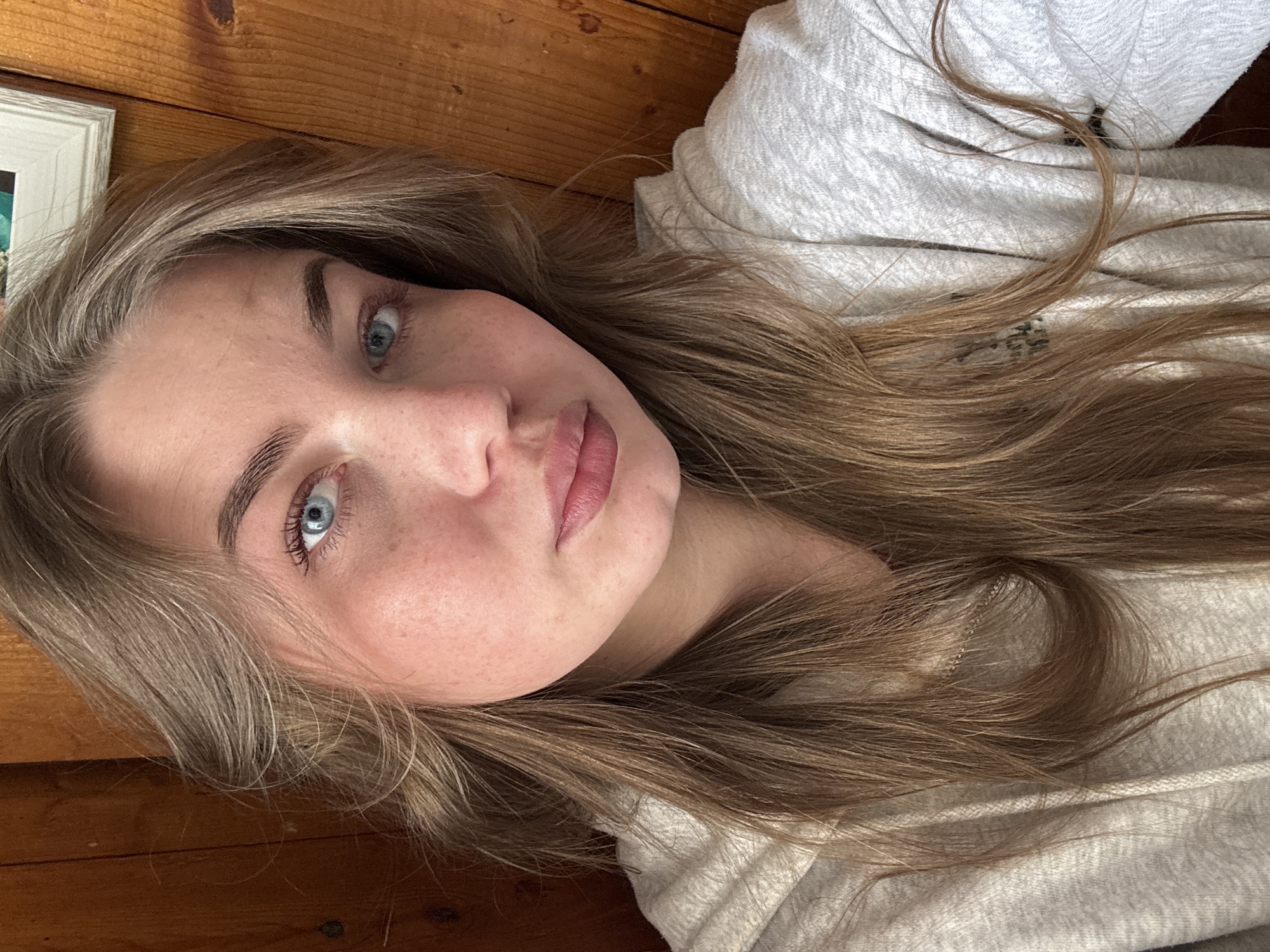 The image is a close-up, diagonal portrait of a young woman with long, light brown hair and striking blue eyes. She is lying down, looking directly towards the viewer with a neutral to slightly relaxed expression. Her lips are tinted with light pink lipstick, and she has visible freckles across her cheeks and nose. Her eyebrows are well-defined.
She is wearing a light grey hooded sweatshirt. Her hair is spread out around her head, with some strands resting on the sweatshirt fabric. The background consists of a warm-toned wooden surface, possibly a floor or wall, against which her head is resting. In the upper left corner of the frame, a small part of a white picture frame is visible.
The lighting appears soft and natural, suggesting an indoor setting. There is no clear indication of the time of day or external weather. On the light grey fabric of her sweatshirt, near her hair, there is some blurry, indistinct text or a graphic, which is not legible. No other specific location cues or text are discernible within the image.
