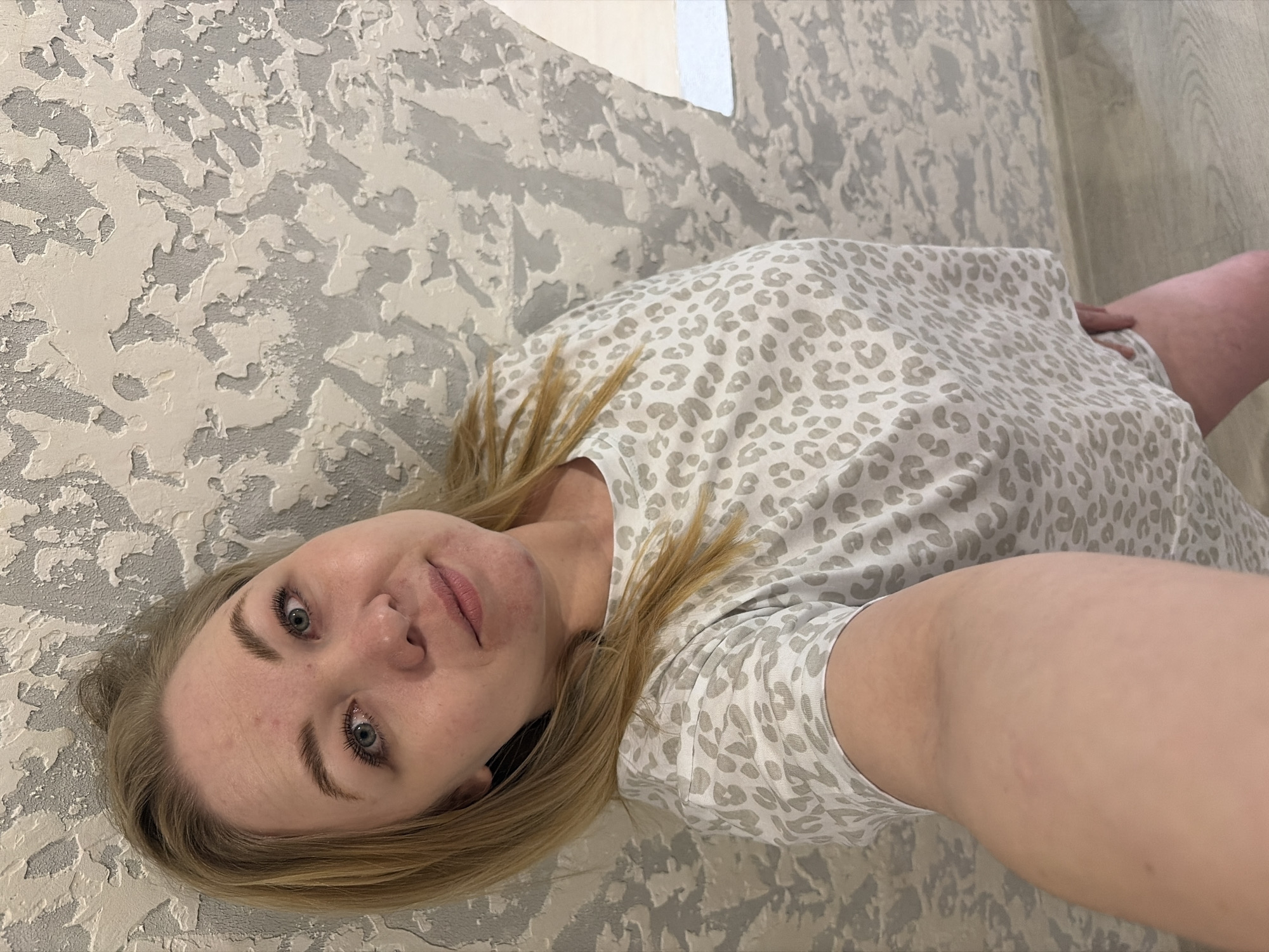 A fair-skinned individual with blonde hair and light-colored eyes (blue-grey) is depicted in a selfie, lying down and looking directly into the camera. Their head is positioned towards the lower left of the frame, with their torso extending towards the upper right. They are wearing a light-colored, short-sleeved top with a subtle grey and beige leopard print pattern. A small black earbud or similar device is partially visible near their left ear. The person has a neutral to slightly pleasant expression with a faint smile.
The background consists of a highly textured wall or surface, predominantly light grey with raised, off-white, abstract patterns. This textured surface occupies the majority of the left and upper parts of the image. To the right of the person, a light-colored wooden floor with visible grain is present. A small, partially visible white object, possibly a piece of paper, is seen near the top edge of the frame. The scene appears to be indoors, likely in a residential setting in Noviy, Russia. There is no visible text in the image.