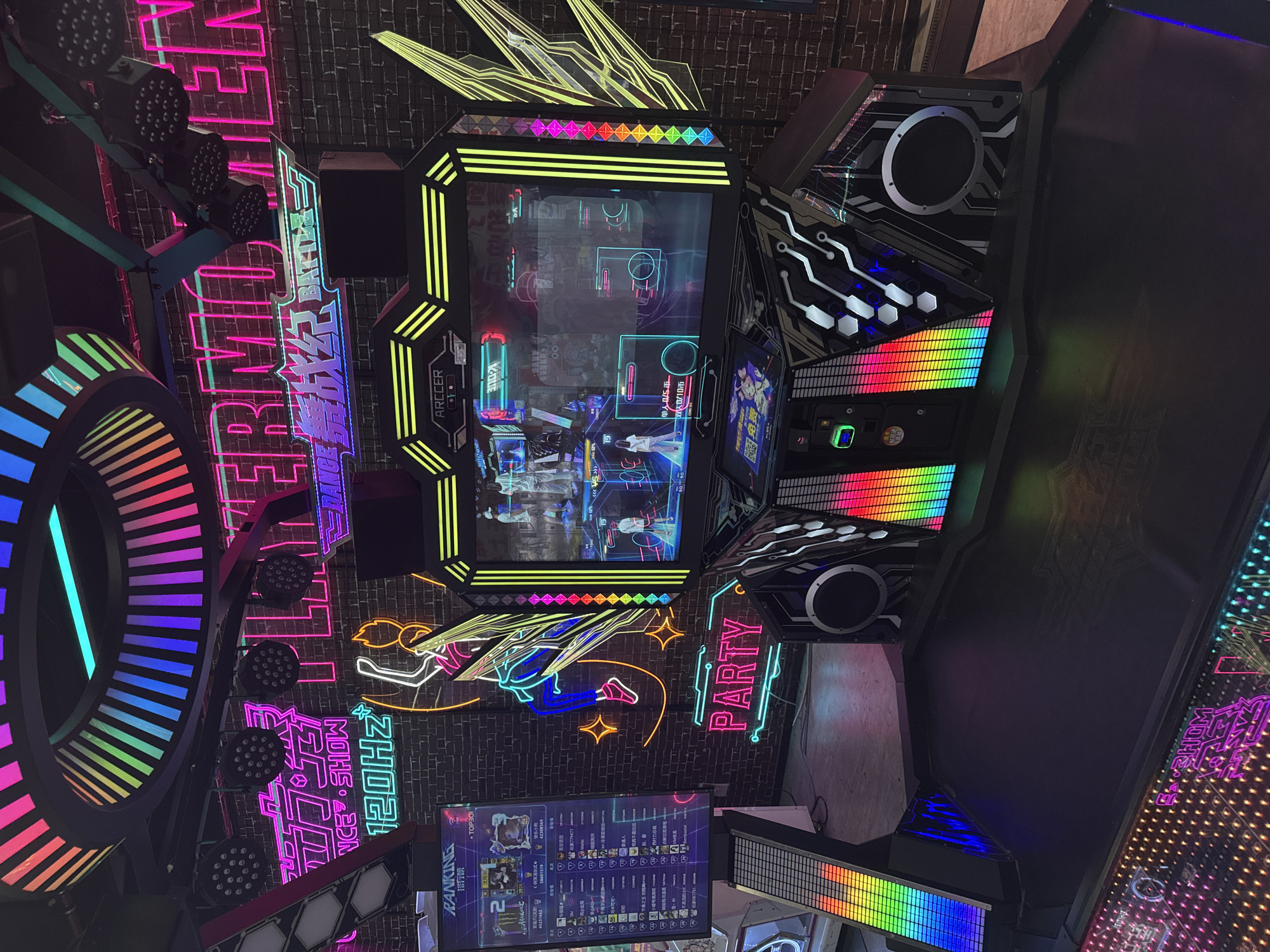 The image depicts an indoor arcade setting, specifically a rhythm game area, within the city of Yiwu, China. The scene is dominated by a large, elaborately lit arcade cabinet designed for a dance or music game. The cabinet features multiple screens displaying colorful graphics and gameplay interfaces, along with numerous LED lights in various colors, including rainbow patterns, pink, purple, and green.
Prominently displayed on the wall behind the arcade cabinet are neon signs that spell out "PLAYERMONSTER" in large, bright pink letters, and other Chinese characters and graphics, including "BATTLE" and "120HZ." There is also a neon outline of a dancing figure. The wall itself appears to be made of brick.
In the foreground, the floor of the arcade is visible, with some areas illuminated by colorful LED lights, forming patterns. The overall atmosphere is vibrant and energetic, characteristic of an entertainment venue. No people are clearly visible in the image, though the presence of an arcade suggests it is a public space. The arcade cabinet itself is the central subject, with its complex design and illuminated components. The context of Yiwu, China, places this arcade within a well-known hub for international trade and commerce, where entertainment venues often cater to a diverse clientele.