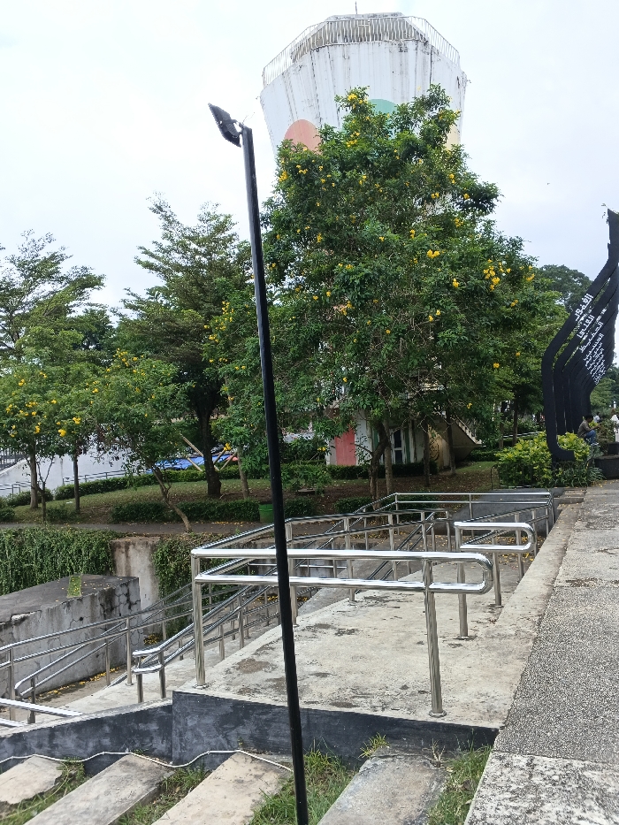 This image depicts a public park in Bien Hoa, Vietnam, featuring the Bien Hoa Water Tower. The scene is captured under an overcast, light grey sky.
In the foreground, a black lamppost with an angular light fixture stands centrally. To its left and extending downwards, rough concrete steps are partially overgrown with grass. In the midground, a multi-level concrete stairway with polished stainless steel handrails descends from the right to a lower level on the left.
The background is rich with vegetation and structures. Multiple green trees, some densely covered with vibrant yellow flowers, are distributed across the landscape. A white retaining wall, partially obscured by green vines, runs horizontally. Further back, small structures or buildings with a red door and a light green door are visible amidst the trees.
To the far right, a tall, black, angular monument or sculpture displays white text, likely Vietnamese script. Two individuals are partially visible near the base of this monument; one is seated, and the other is partially obscured behind them.
Dominating the upper background is the Bien Hoa Water Tower (Tháp Nước Biên Hòa), a tall, white, octagonal structure with a perimeter railing at its top. Its side features three large colored circular designs in orange, light green, and a darker green or blue. The ground throughout the park area is covered with grass and manicured shrubs. A green trash bin is positioned under a tree on the left side of the large, yellow-flowered tree. No explicit actions or interactions beyond static presence are discernible among the individuals.