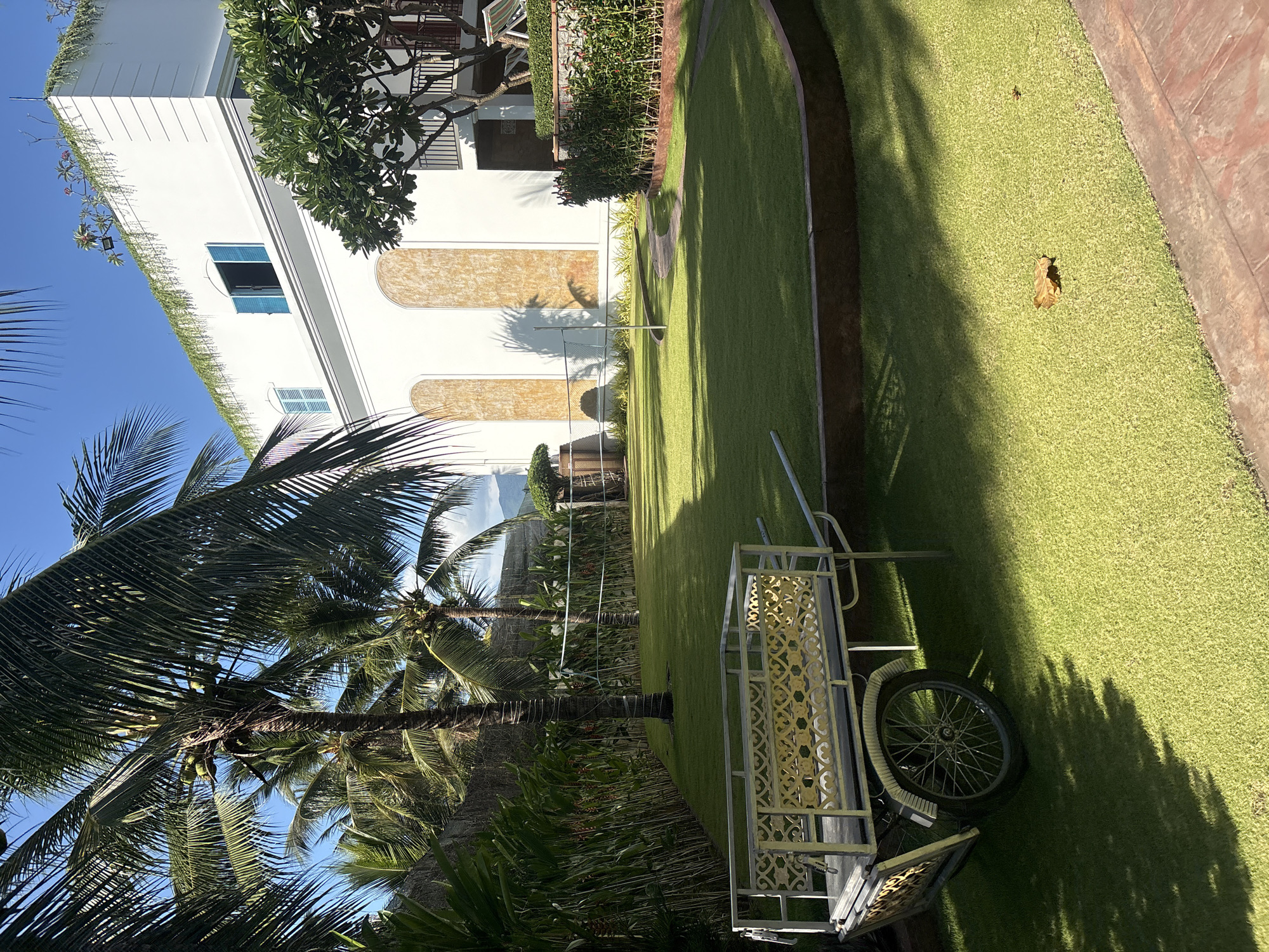 The scene is a sunny outdoor area in Bac Nha Trang Ward, Vietnam, as indicated by the architecture and vegetation. A white, multi-story building with distinct architectural features is present, partially obscured by tropical trees. The foreground includes palm trees with coconuts. A decorative metal cart is positioned on a well-maintained green lawn. The lighting casts strong shadows across the grass.