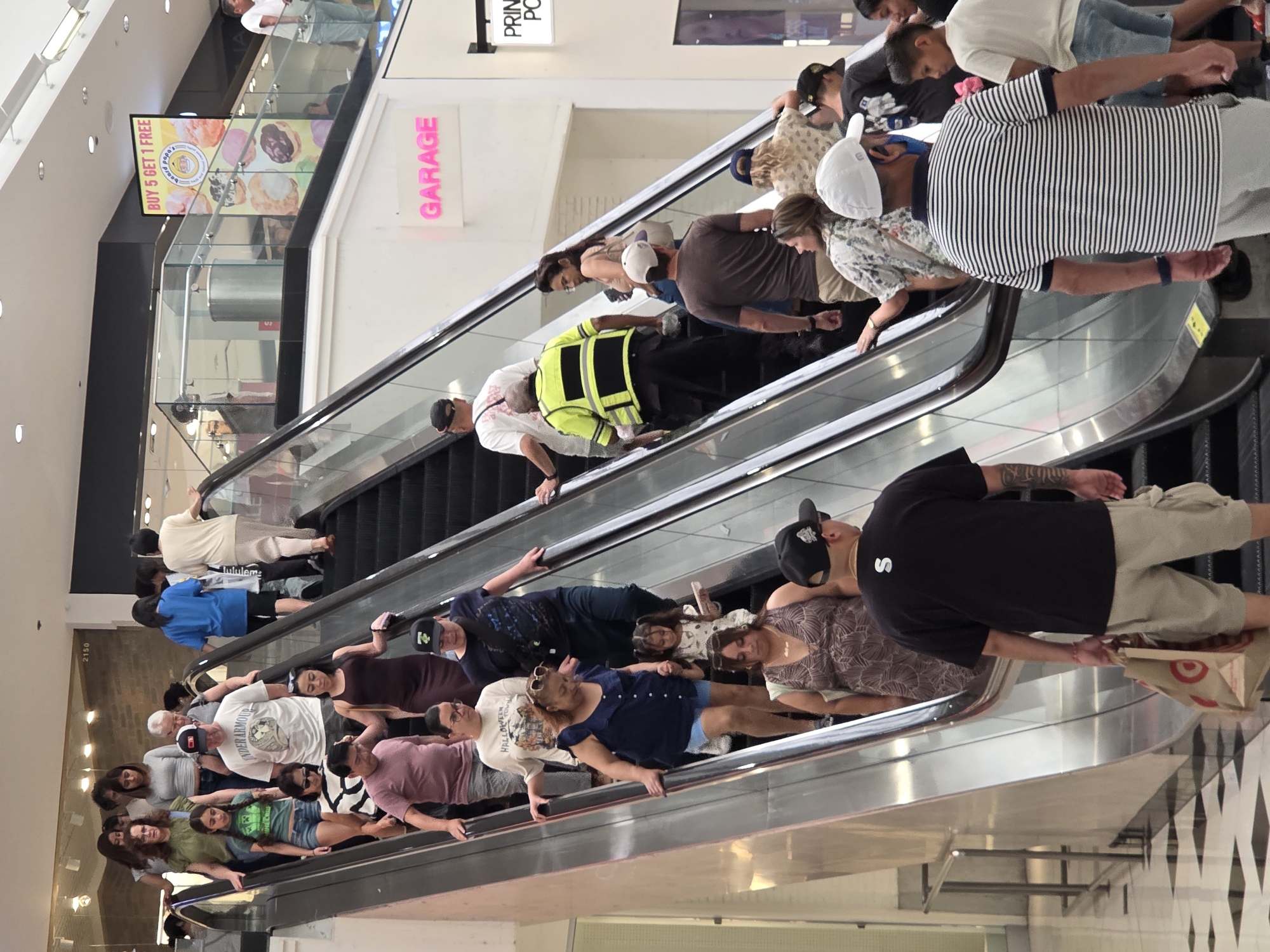 In Glendale, United States of America, a group of people are ascending an escalator in a shopping mall. The escalator is metallic with black steps. Several individuals are visible on the escalator, with some holding onto the handrail. A sign displaying "BUY 5 GET 1 FREE" with images of donuts is visible above and to the right of the escalator. Another sign with the word "GARAGE" in pink neon letters is also visible. The mall's flooring is checkered black and white. Some people are carrying shopping bags. The overall scene depicts everyday activity within a commercial establishment.