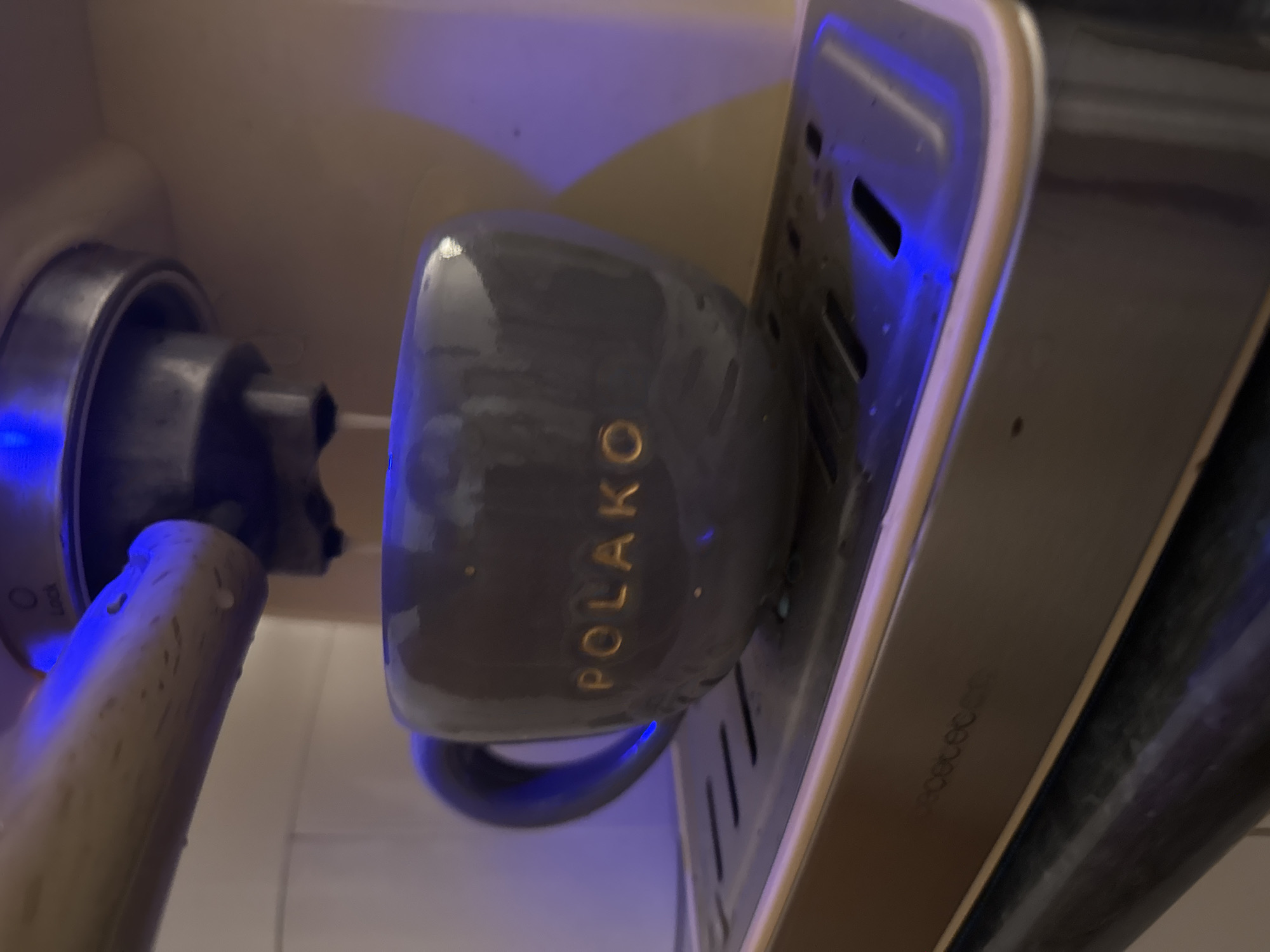 The image presents a close-up, rotated view of a coffee preparation area, likely within a kitchen or cafe setting in Zankovići, Montenegro. A grey, glossy ceramic coffee mug is the central subject, resting horizontally on a metallic drip tray or cup holder that features slotted drainage. Prominently displayed on the mug in gold lettering is the word "POLAKO."
To the left of the mug, a metallic component resembling a portafilter from an espresso machine is visible, with a light-colored handle extending downwards and to the left. The handle appears to have water droplets or condensation on its surface. Part of a dial or mechanism is also visible near the portafilter, with the faint text "LOCK" discernible.
The scene is dramatically illuminated by a strong, artificial blue light, which reflects intensely off the glossy surfaces of the mug and the metallic elements of the machine, creating a cool, almost futuristic ambiance. This lighting suggests either low ambient light conditions or a deliberate aesthetic choice. The background is a soft, indistinct beige or off-white wall. No people are present, but the arrangement indicates the potential for, or a recent instance of, beverage preparation.