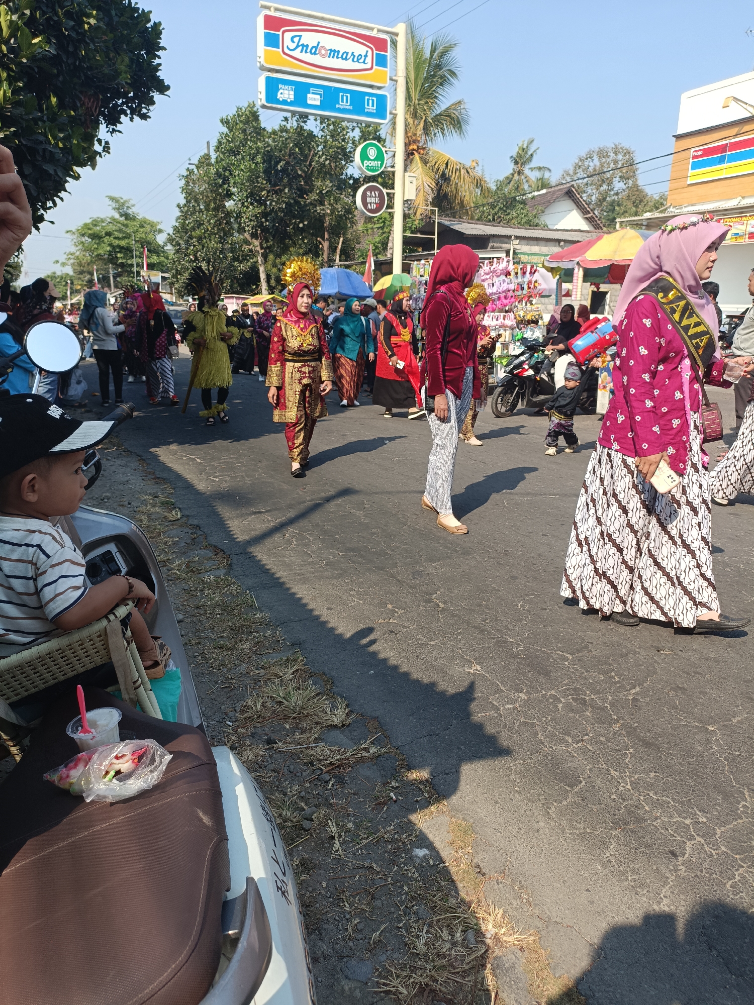 In Jatitengah, Indonesia, a parade is in progress on a sunny day. The street is lined with trees and buildings, including an Indomaret convenience store. People are walking in the street, many of them wearing traditional Indonesian clothing. Some are dressed in elaborate costumes, while others wear more casual attire. A child in a black cap sits on a scooter in the foreground, looking to the side. Food items are visible on the scooter's seat. A woman in a pink hijab and a magenta dress with a sash that reads "JAWA" is walking away from the camera. Another woman in a red outfit and a gold headpiece is walking toward the camera. The atmosphere appears festive, with a sense of community and cultural celebration.
