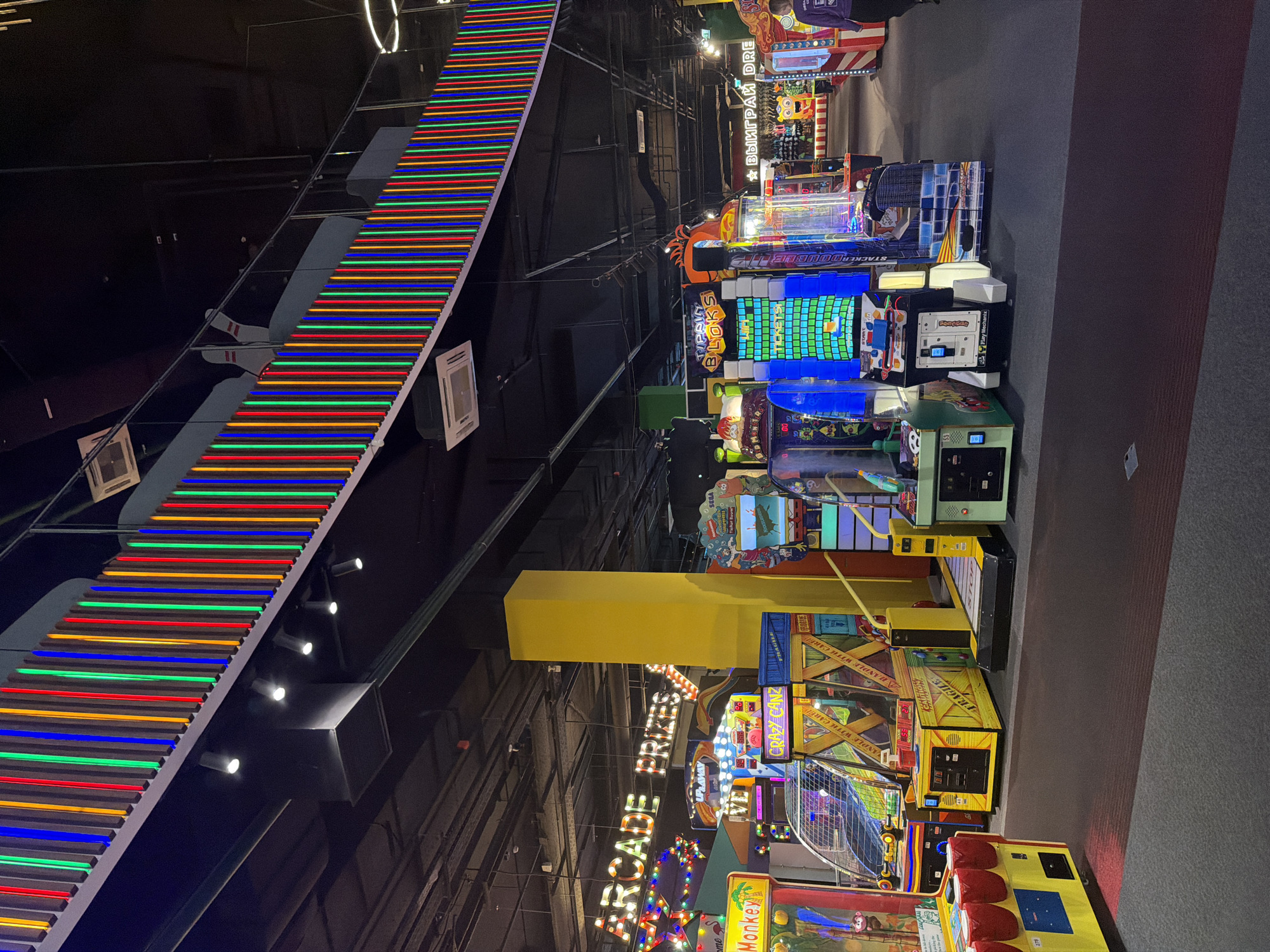An indoor amusement arcade is shown, with rows of colorful arcade machines and neon lights. A prominent yellow pillar stands in the center, and signage for "ARCADE PRIZES" is visible. Various game cabinets are visible, including ones labeled "Crazy Canz" and "Tip Top Blocks". A person in a purple jacket is walking away from the camera in the background on the right. The overall scene is brightly lit and filled with the visual stimuli typical of an entertainment venue.