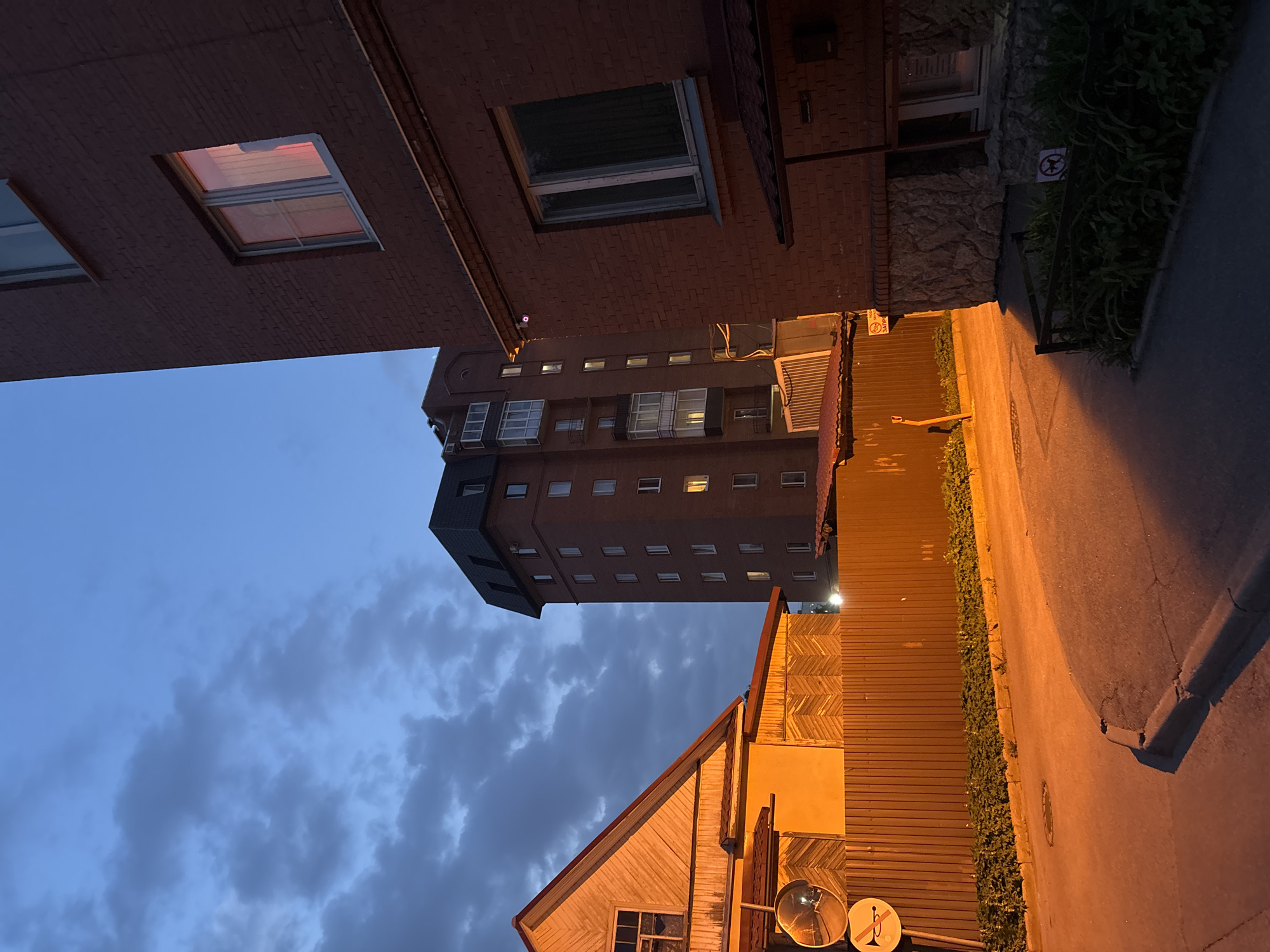 The image depicts a street scene in Saint Petersburg, Russia. The view is oriented upward, showing a mix of buildings against a blue, cloudy sky. A tall, red brick apartment building is centrally placed, with numerous windows visible. Adjacent to the apartment building are older, shorter structures: one with visible windows and a wooden exterior, and another with red brick and an attic window. The street is narrow, bordered by a fence and foliage. The lighting suggests either early evening or late dusk, with a warm glow emanating from the street lamps.