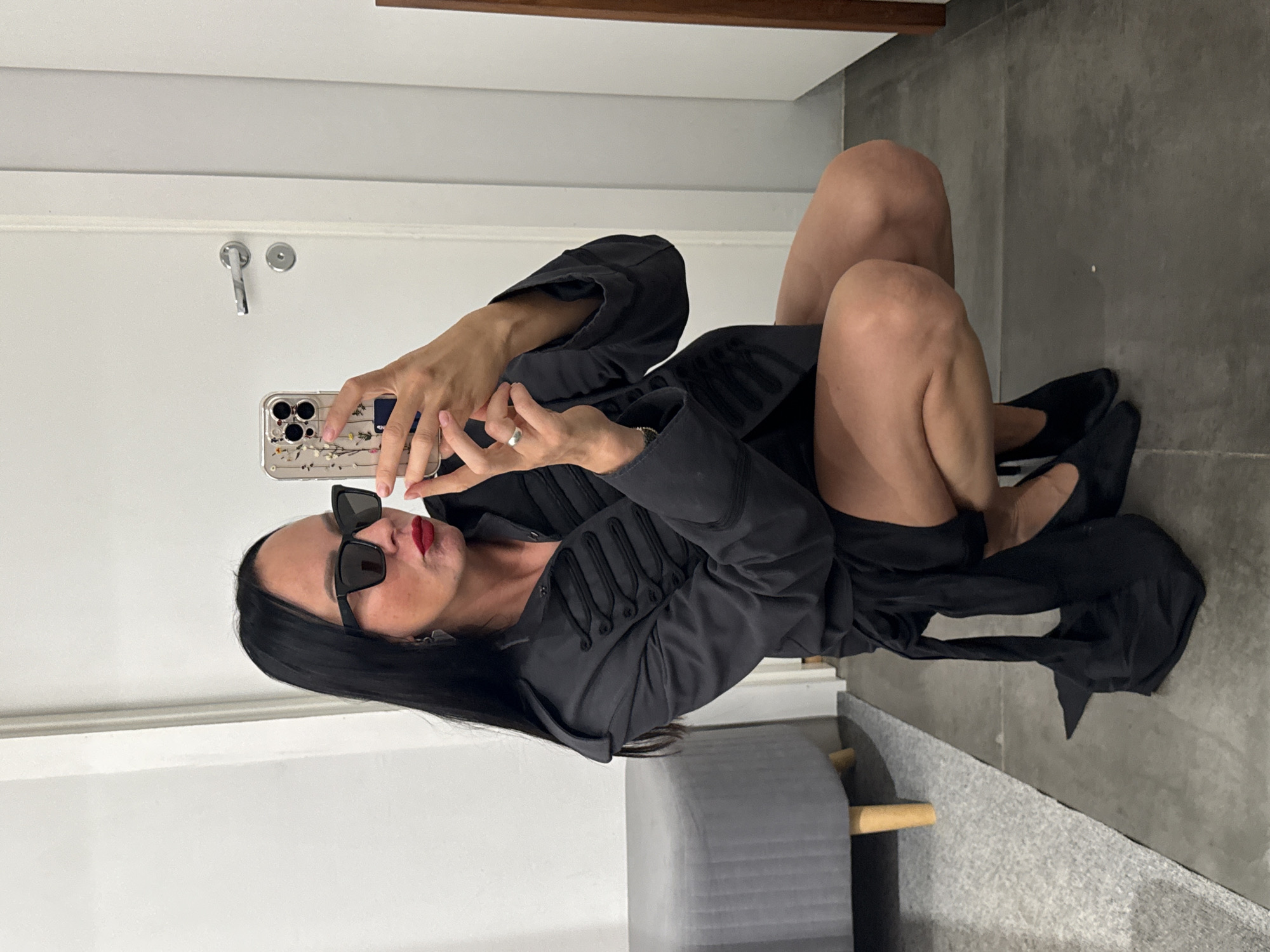 A woman with black hair, wearing sunglasses, a dark jacket with ornate buttons, and a short black dress, is squatting in front of a full-length mirror and taking a selfie with her phone. The jacket has a high collar and epaulets. She is wearing black pointed-toe shoes. A gray upholstered bench with wooden legs is visible to the left of the woman, and a white door with a silver handle is behind her. The floor appears to be tiled. The image does not provide any information that would indicate it was taken in Chaikovsky, Russia.