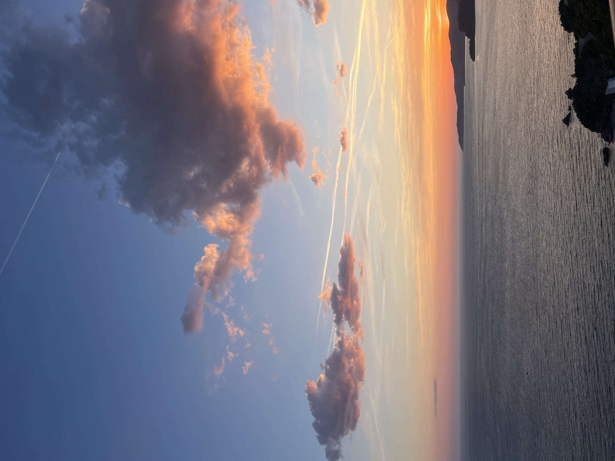 Captured at Sveti Stefan, Montenegro, the image presents an expanse of calm sea, its surface subtly textured by wind. Adjacent to the sea, a rugged, verdant coastline is visible. The sky transitions from a clear, deep blue to a soft, gradient sunset of oranges and yellows near the horizon. Scattered throughout the sky are billowy clouds, illuminated by the setting sun, casting a warm, rosy hue on their surfaces. Two aircraft contrails traverse the sky, adding linear elements to the composition. No people are present in the image.