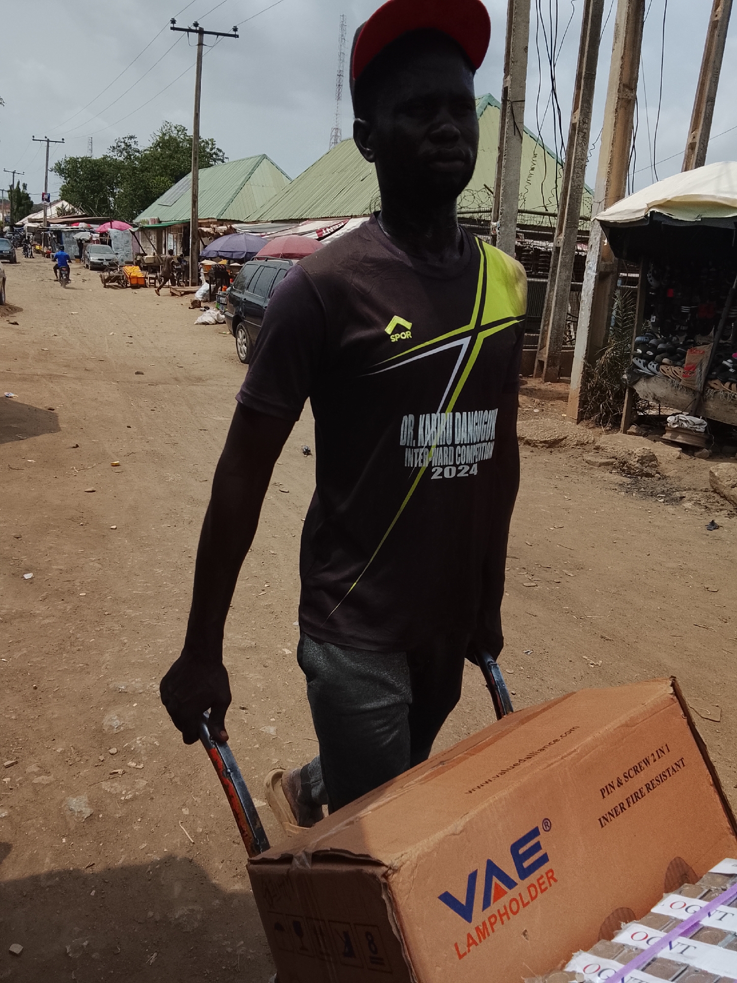 A person is pulling a hand truck loaded with boxes. The boxes are labeled "VAE LAMPHOLDER" and "PIN & SCREW 2 IN 1 INNER FIRE RESISTANT". The person is wearing a t-shirt that says "SPOR" and "DR. KABIRU DANGUGU INTER-WARD COMPETITION 2024". The person is wearing a red hat. The location appears to be a street market in Abuja, Nigeria.