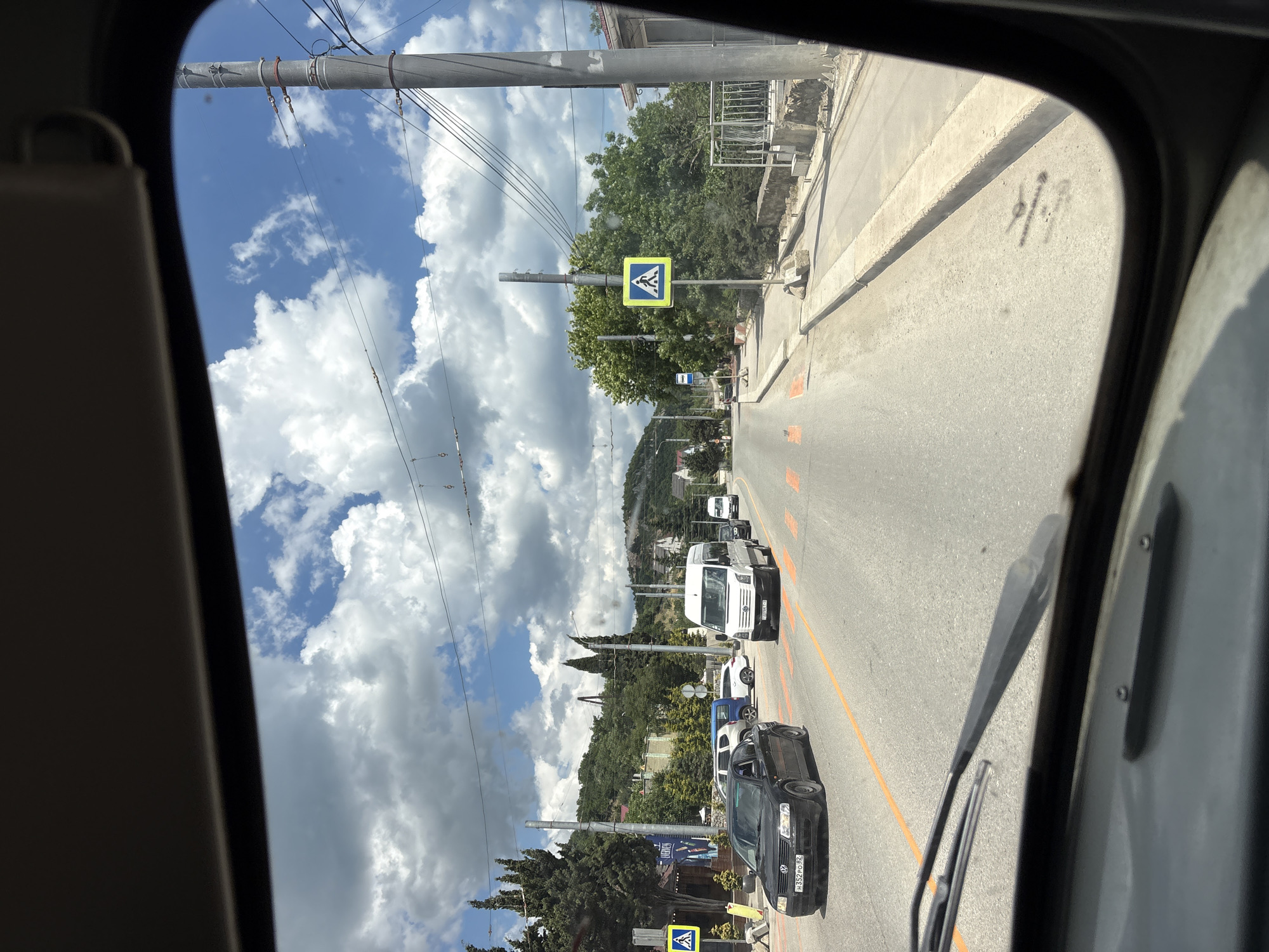 The image is taken from the interior of a vehicle, looking through the windshield. It shows a sunny day with scattered clouds. The view is of a road with several cars and vans. There is a pedestrian crossing with orange markings on the road. A yellow pedestrian crossing sign is visible on the right side of the road. Trees and buildings are visible in the background, with a hilly landscape behind them. The location is identified as Kiparisnoe, Ukraine.