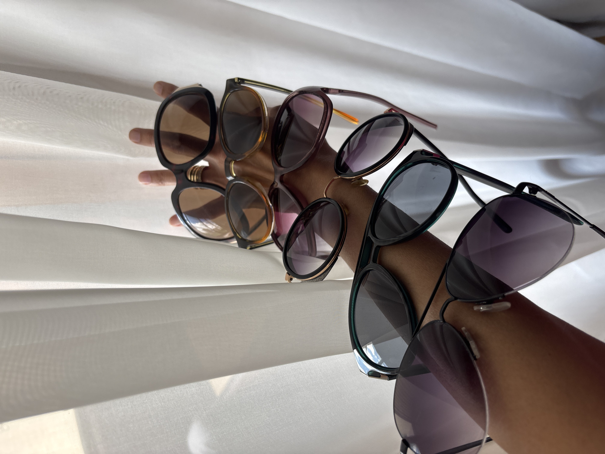 A hand holds several pairs of sunglasses in front of a white curtain. The sunglasses vary in style, color, and lens tint. The hand is light brown in complexion. The curtain is thin, allowing some light to pass through.
There is no indication of the location being in Minsk, Belarus, or any other specific location.