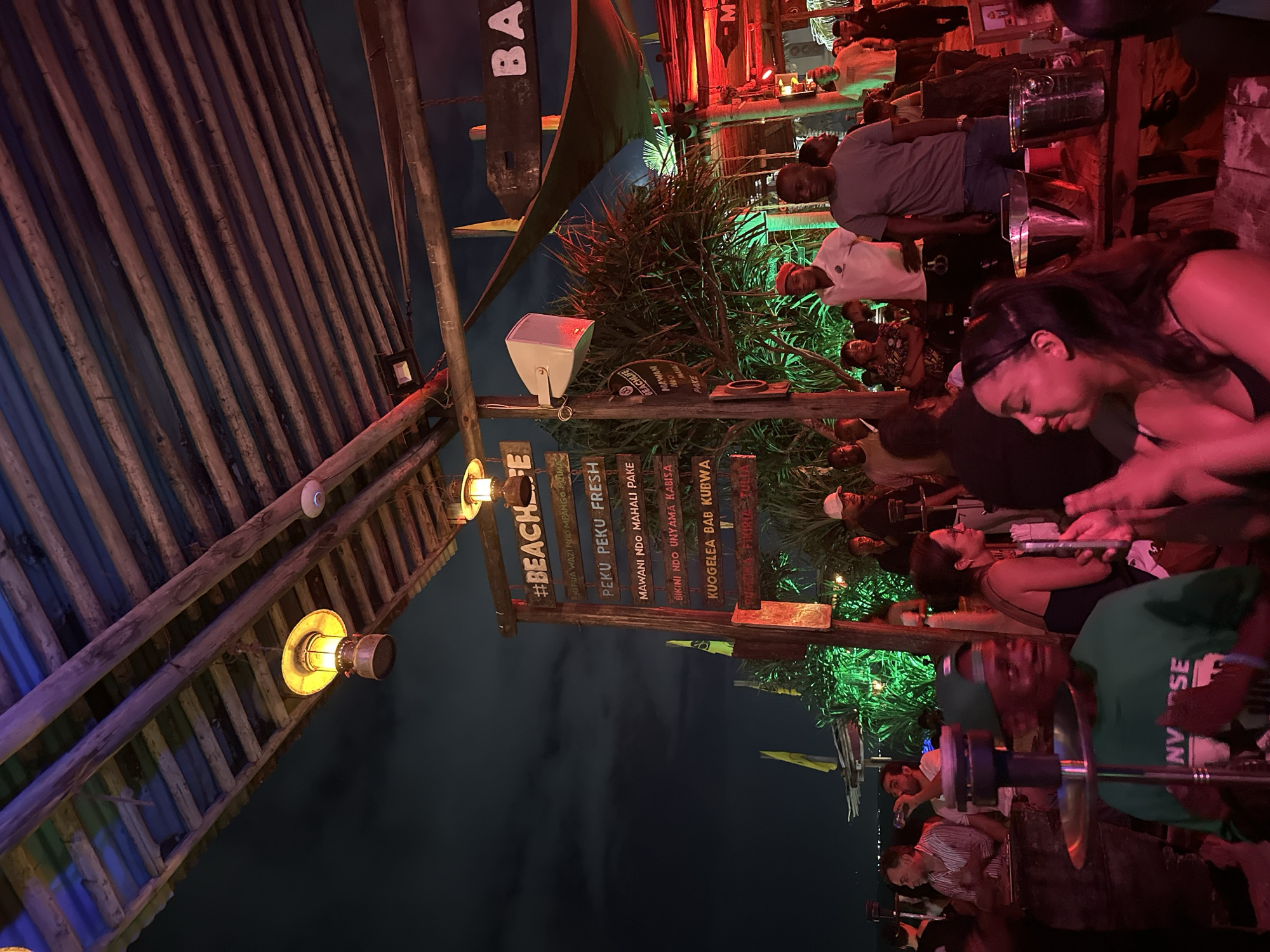 The image depicts a busy outdoor bar scene at night. People are gathered around tables, some appearing to be smoking from hookahs. Wooden structures and thatched roofs create a rustic aesthetic. A prominent sign reads "#BEACHE" along with other phrases in Swahili, suggesting a beachside location in a Swahili-speaking region. The lighting is dim with colored lights, particularly red and green, contributing to the ambiance. The environment is lively, suggesting a social gathering in the city of Unknown, Unknown.