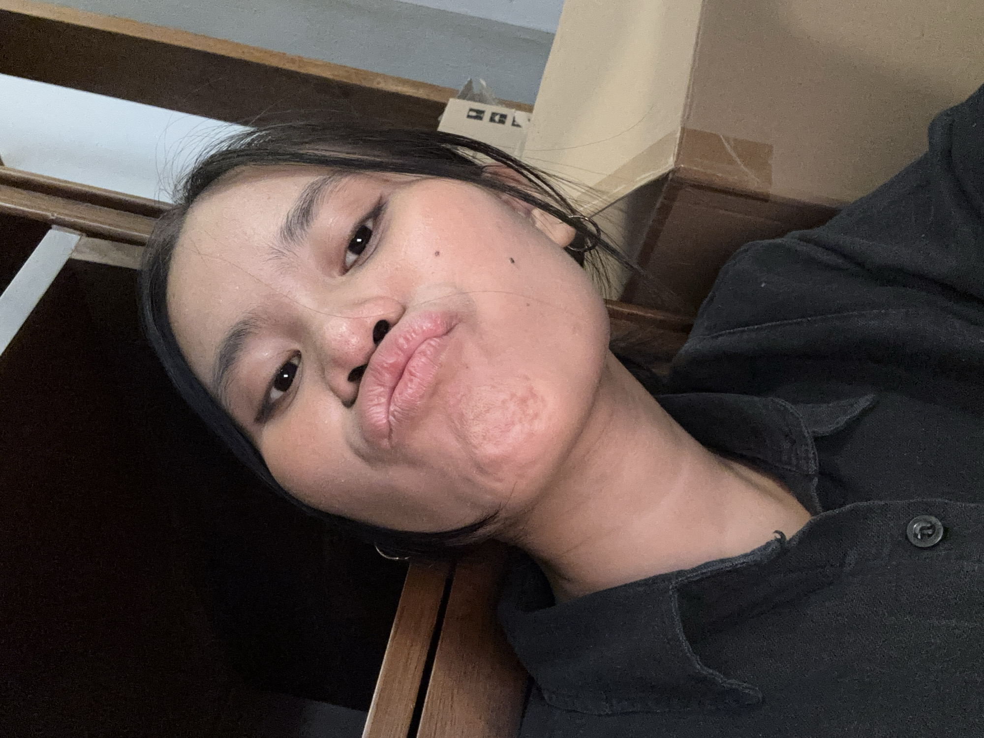 A young woman with dark hair and fair skin is taking a selfie. She is wearing a black collared shirt. Her lips are pursed, and she is looking directly at the camera. The background is cluttered with wooden furniture and cardboard boxes, suggesting an indoor, possibly residential setting. No specific location in South Jakarta, Indonesia can be determined from the image.