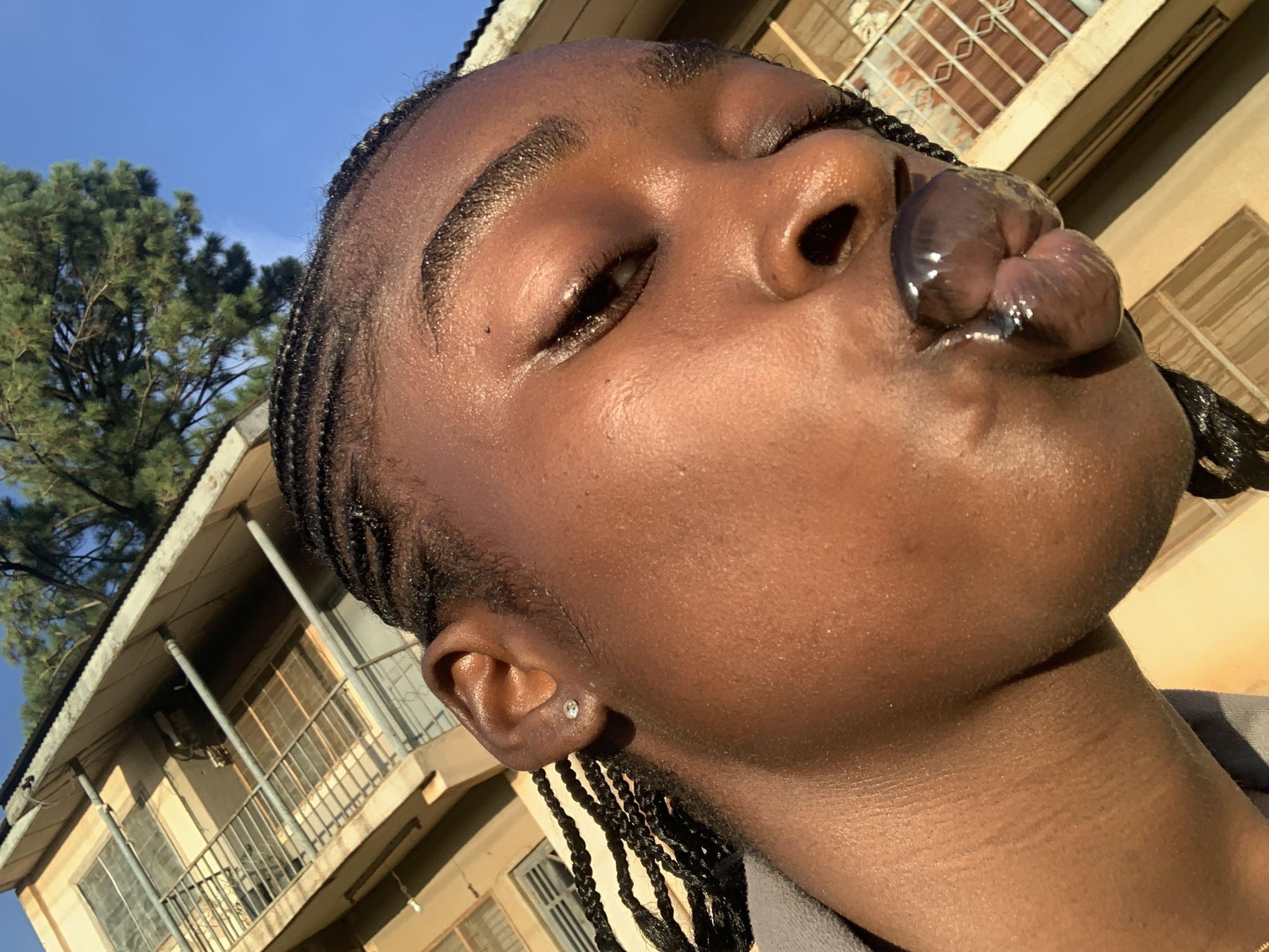 A close-up, head-and-shoulders selfie shows a young Black woman in a gray top, her face turned slightly to her right, with her lips pursed as if blowing a kiss. Her dark braided hair frames her face. She wears a small, sparkling stud earring in her visible left ear. The background features a light-colored building with balconies and windows, bathed in sunlight, against a clear blue sky. A green tree is visible in the upper left corner. The overall scene suggests an outdoor setting during the daytime. The image was taken in the city of Rafin Par, Nigeria.