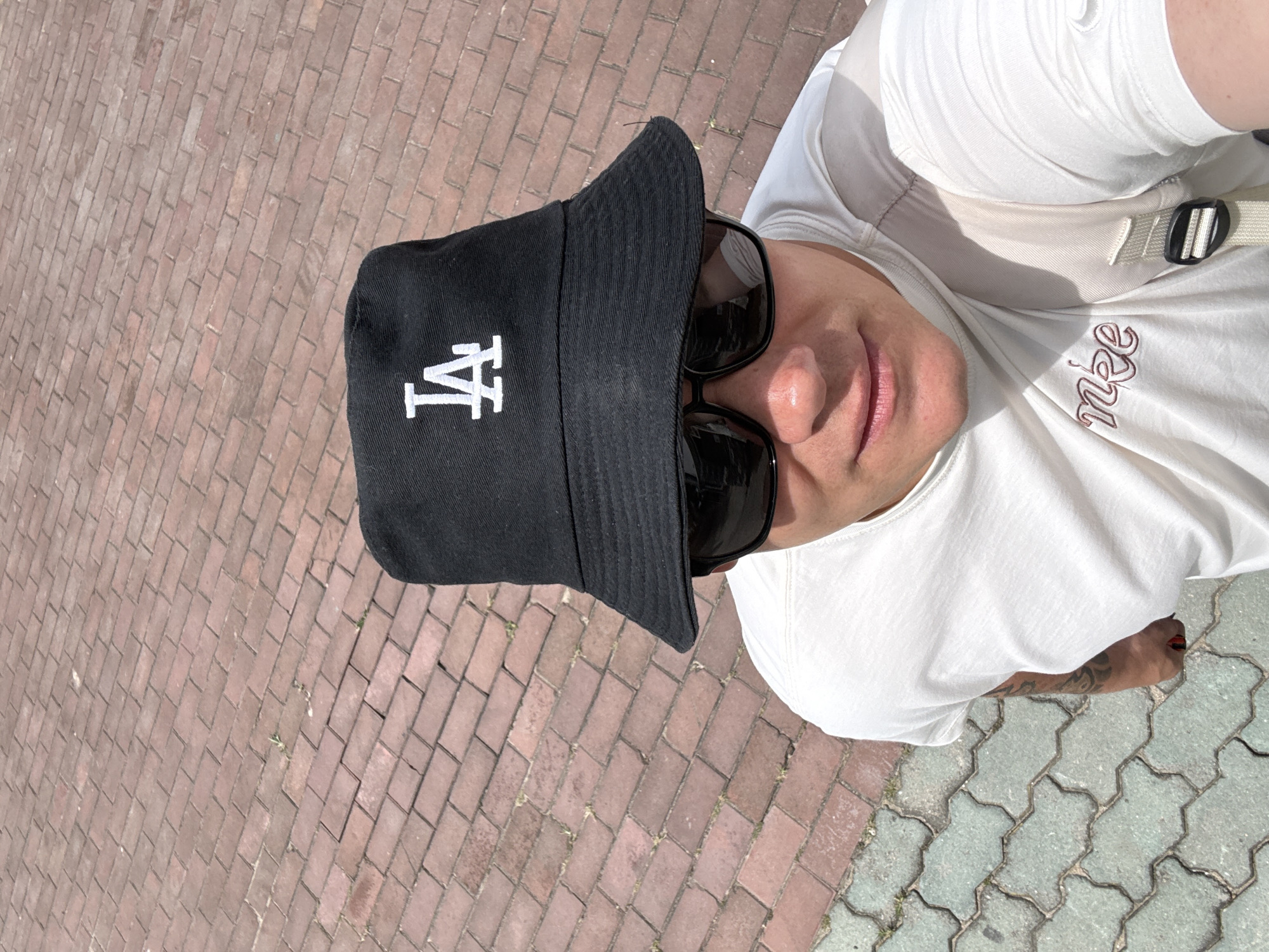 A person wearing a white t-shirt, black bucket hat with "LA" logo, and sunglasses is posing for a selfie outdoors on a sunny day. The person has tattoos on their arm. The ground consists of red brick paving stones and some lighter colored interlocking pavers. The background is blurred. The location is not identifiable as Qinhuangdao, China.