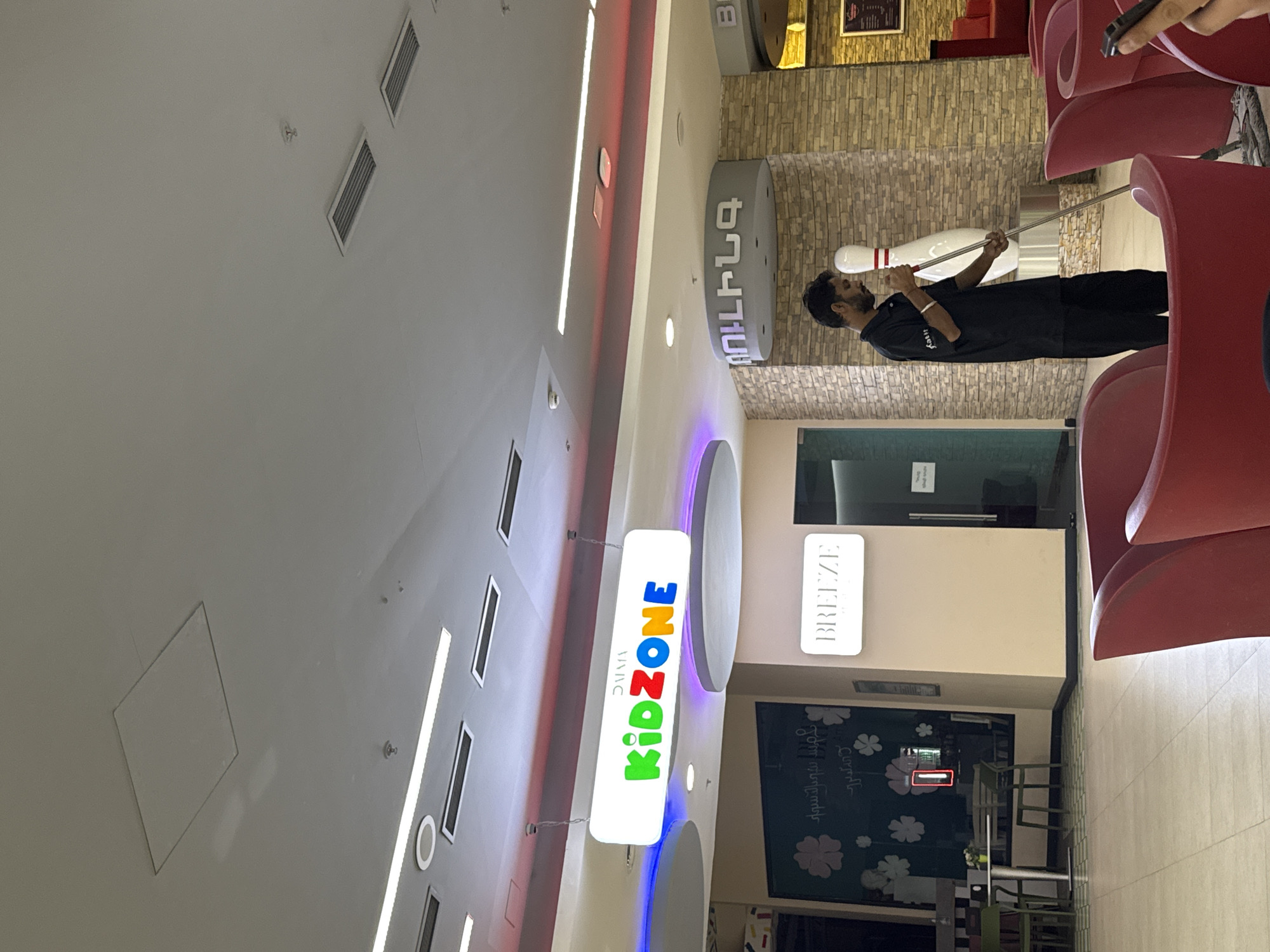 The image shows the interior of a shopping mall or entertainment center in Yerevan, Armenia. Signage indicates the presence of "KidZone" and "Breeze" within the venue. A man in a black uniform appears to be engaged in maintenance or repair work, holding a bowling pin on a rod. Nearby, red seating is visible. The location has modern interior design elements, including illuminated signs, linear lighting, and ceiling ventilation.