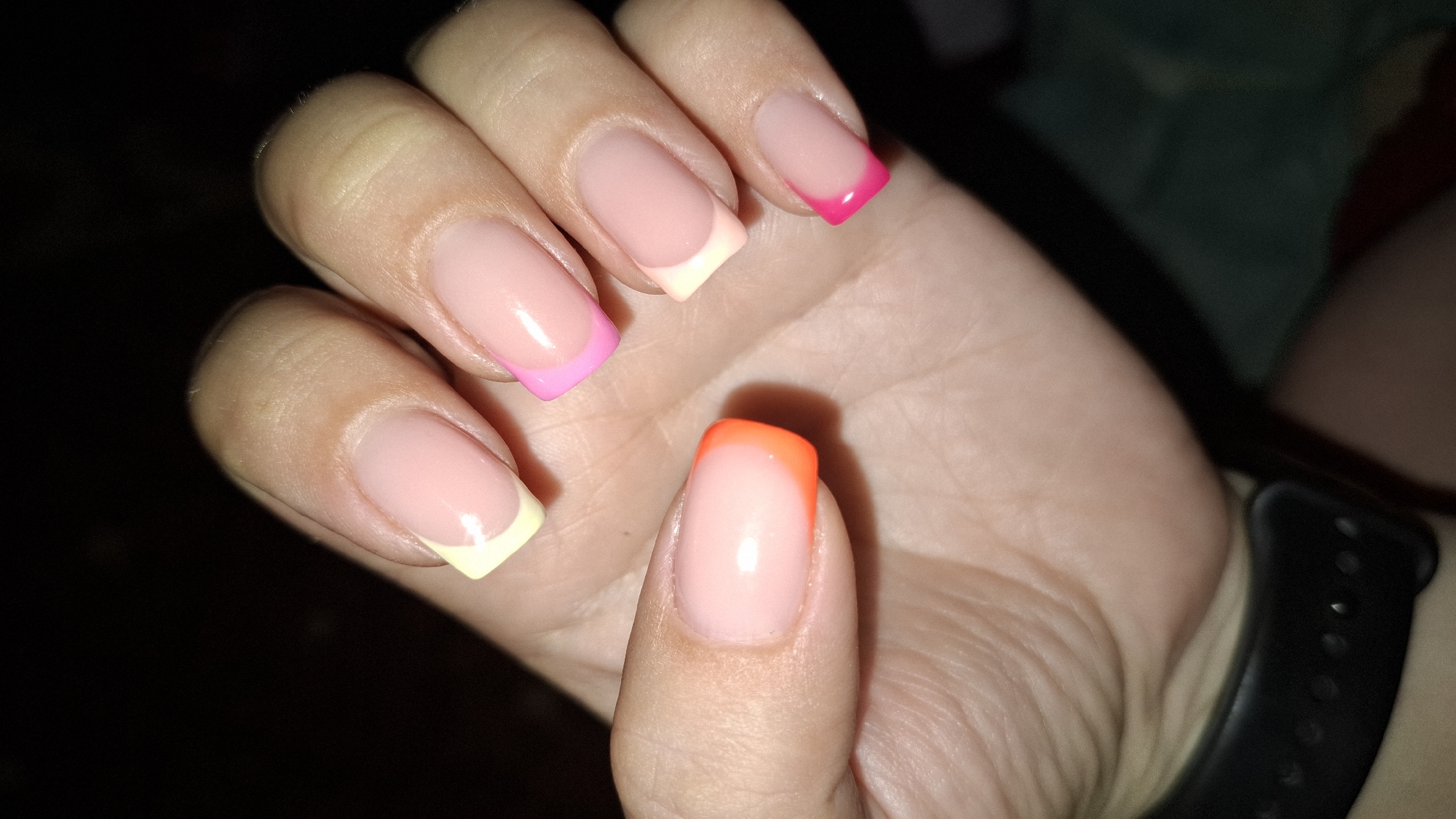 A close-up, dimly lit, over-the-shoulder shot shows a person's left hand with brightly colored French manicured nails. The nails have a sheer pink base with vibrant tips in yellow, pink, orange, and hot pink. The hand is positioned against a dark background, possibly a night scene in Шұбарсу, Kazakhstan, with the blurred outline of a dark smartwatch band visible on the wrist.