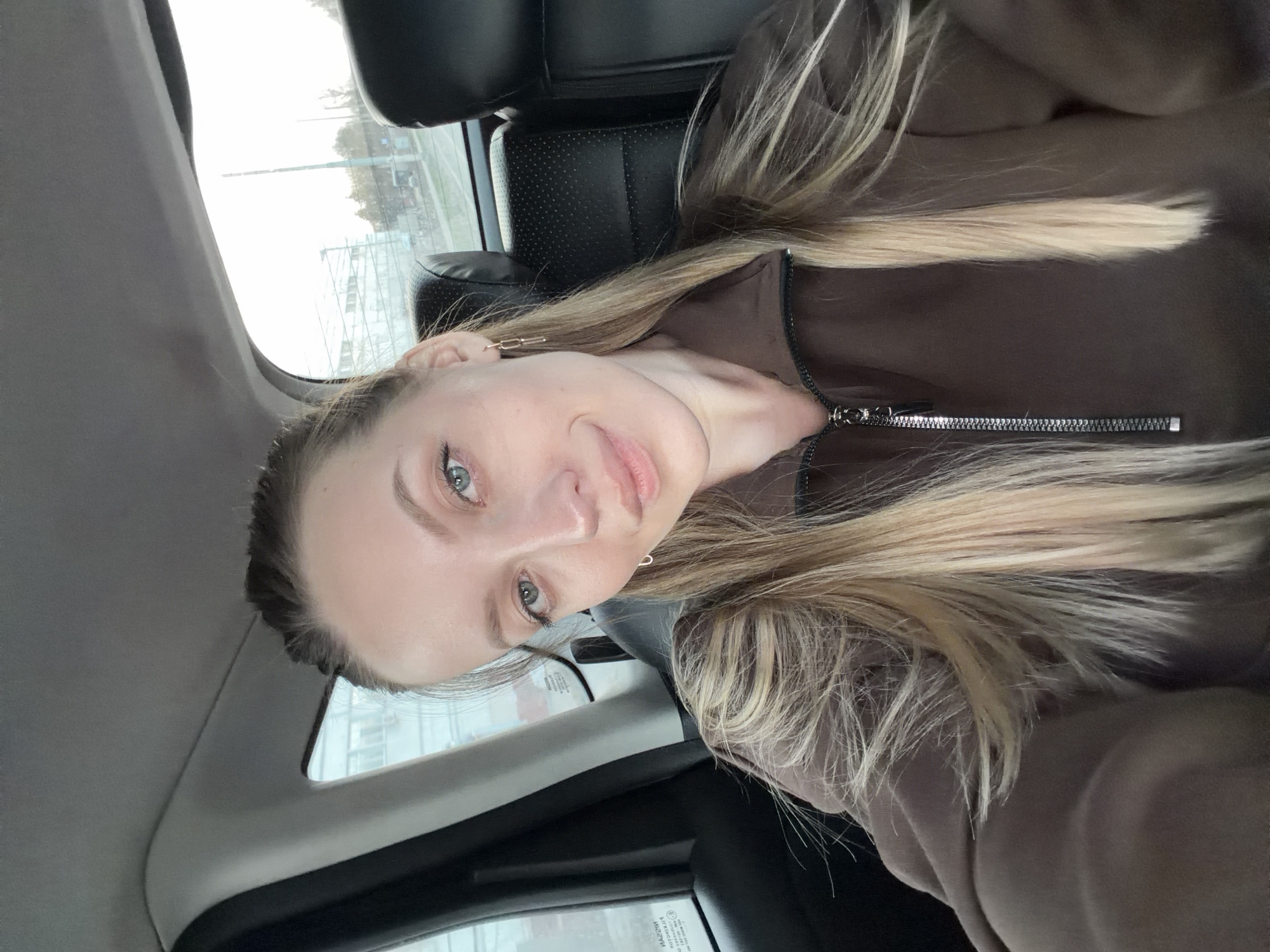 A woman is pictured inside a car. The car is parked on a street in Klin, Russia. The woman has blonde hair, fair skin, and light-colored eyes. She is wearing a brown jacket. The interior of the car consists of black seats and trim. Outside the window is a view of a city street with buildings and streetlights. The weather appears overcast.