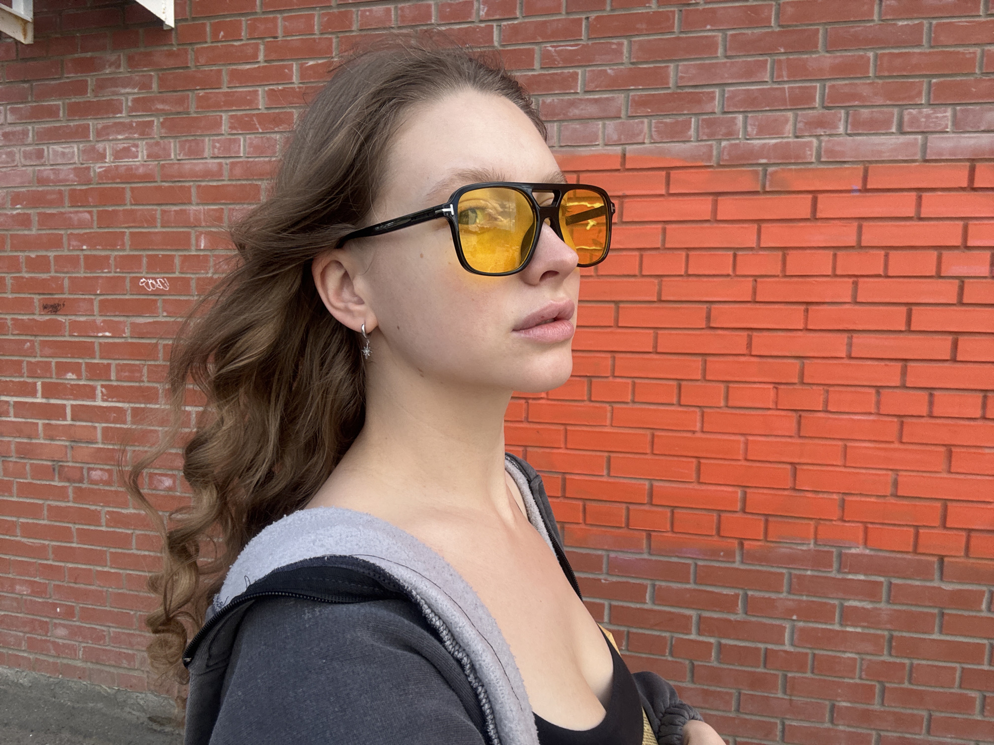 A young woman with light brown hair styled in loose waves stands outdoors in front of a brick wall, her body angled to the right and her head turned to the left. She wears large, black-framed sunglasses with yellow lenses, a gray zip-up hoodie over a black tank top, and a small silver star-shaped earring in her left ear. The brick wall behind her is a mix of reddish-brown and bright orange bricks, with some faint graffiti visible on the darker section. The lighting suggests it is daytime, with shadows cast on the wall. The woman appears to be walking or posing for a photo. The scene is located in the city of Izhevsk, Russia.
