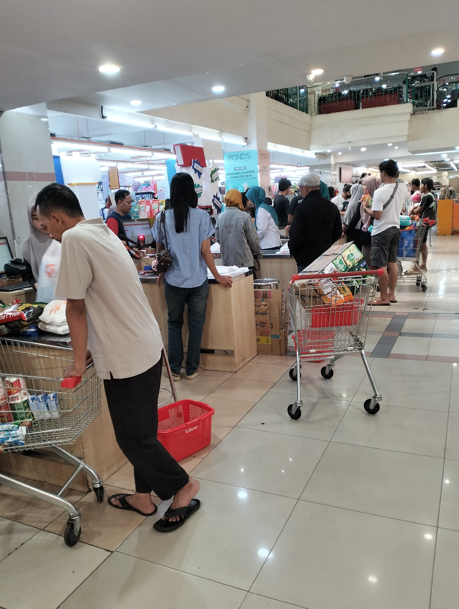 The image captures a scene inside a brightly lit supermarket in Ungaran, Indonesia, likely near a checkout area. A man in a light-colored polo shirt and dark pants is standing at a checkout counter, leaning on his shopping cart, which is partially filled with groceries and a red basket. His posture suggests he is waiting or interacting with a cashier, though the cashier is not clearly visible.
In the background, several other shoppers are present, some waiting in line, others browsing. A woman in a blue striped shirt and jeans is also at a checkout counter, facing away from the camera. Another woman in a light hijab and a grey jacket is standing near a checkout counter on the left.
The environment appears to be a typical supermarket aisle, with shelves stocked with products visible in the distance. The floor is tiled, reflecting the overhead lighting. Shopping carts and baskets are scattered throughout the area, indicating active customer activity. The overall impression is one of a busy retail environment during operating hours.
