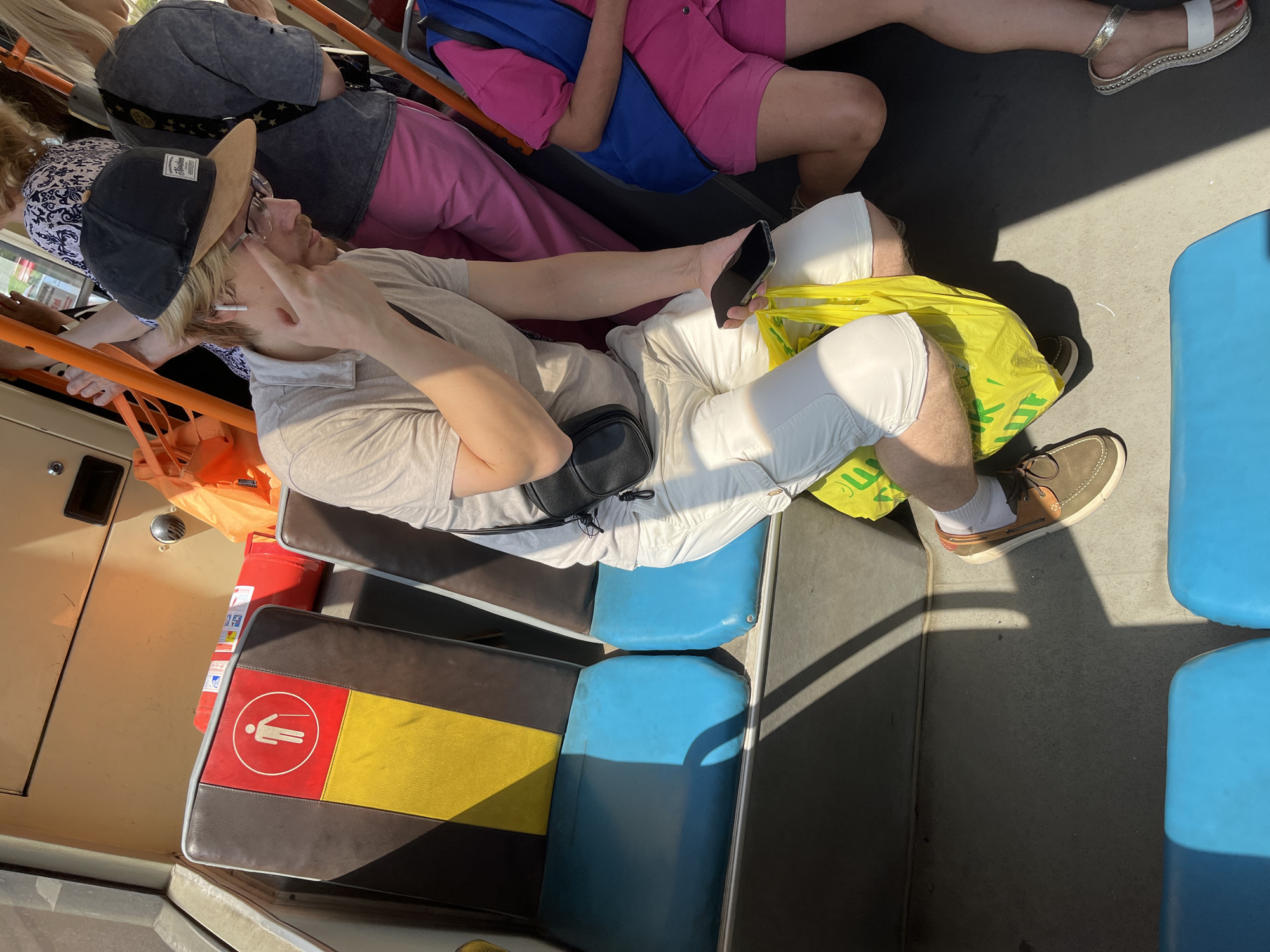 The image shows an interior view of a public transport vehicle, likely a bus, with several passengers. In the foreground, a person wearing white shorts, a light-colored polo shirt, and a baseball cap is seated, holding a smartphone and a bright yellow plastic bag. They are also wearing brown and white boat shoes. Next to them, another person is visible from the knees down, wearing pink shorts and a blue vest, with one foot in a white sandal. In the background, other passengers are partially visible, including one wearing a blue and white patterned top and another in a grey t-shirt. The seats are blue and brown with yellow accents. A red fire extinguisher is also visible on a seat. The lighting suggests it is daytime, with strong shadows cast across the floor. The scene is consistent with a typical public transportation environment in Minsk, Belarus, given the general appearance of the vehicle and passengers.