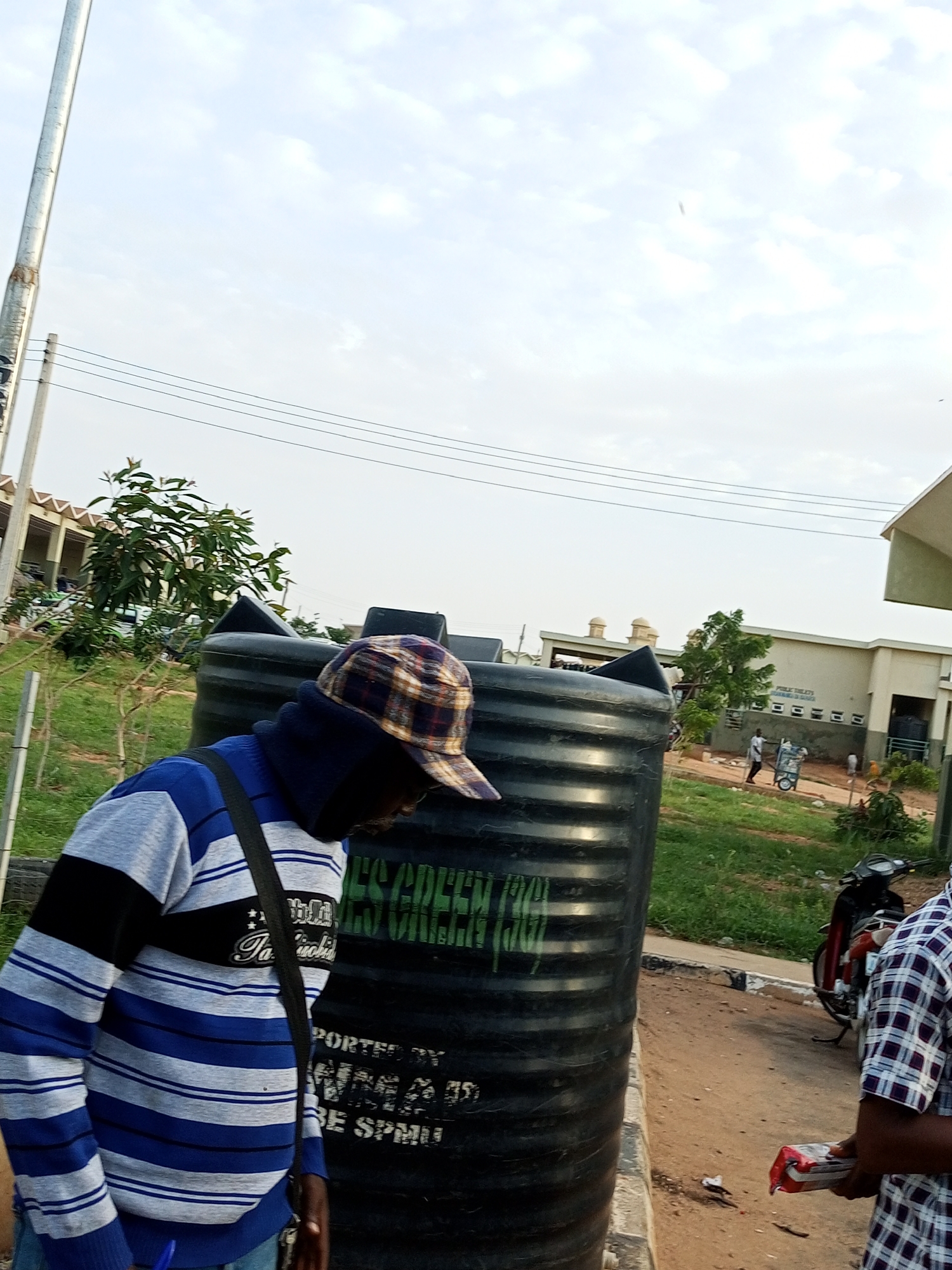 The image depicts an outdoor scene in Gombe, Nigeria, with a focus on a large black water tank situated in the foreground. A man wearing a blue, grey, and black striped sweater and a plaid cap is standing to the left of the tank, looking downwards. He has a dark strap from a bag or satchel across his chest. The water tank has text printed on its side, including "PUBLIC TOILETS" and "GREEN". In the background, there are buildings with domed roofs and a section of a beige structure with "PUBLIC TOILETS" written above a row of square openings. Several individuals are visible in the mid-ground, including one man pushing a cart. A red and black motorcycle is parked to the right of the water tank. The environment appears to be a semi-arid area, with dry soil and sparse green vegetation. The sky is overcast with scattered clouds.