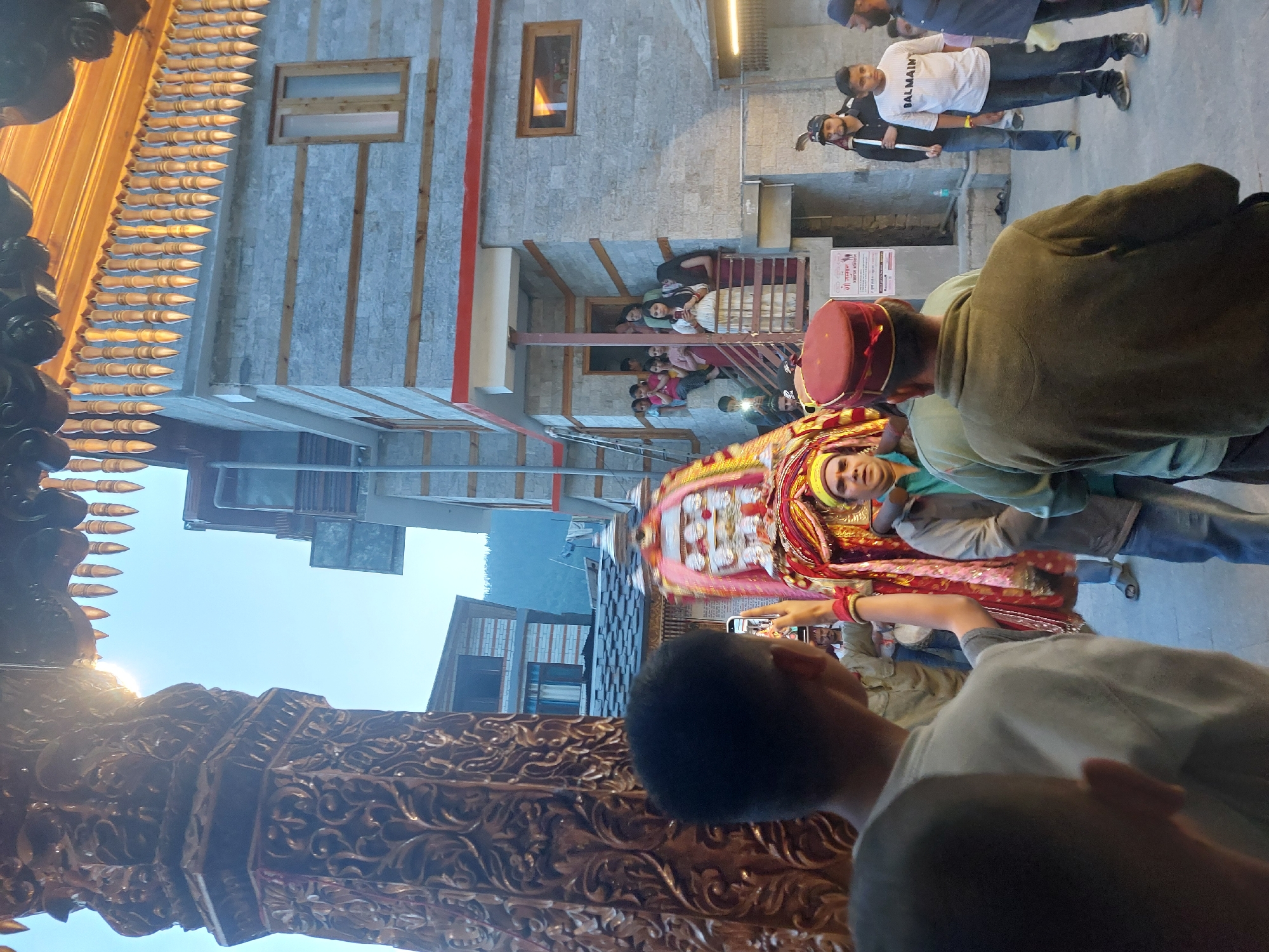 In Vashisht, India, a procession is underway. A group of people are carrying a decorated palanquin, adorned with colorful fabrics and religious imagery, through a street. The palanquin is supported by poles and appears to be carried by multiple individuals. Onlookers line the street, some watching from balconies of traditional stone buildings, while others observe from ground level. The architecture of the buildings features stone construction with wooden accents, characteristic of the region. The sun is visible, casting light on the scene, and the sky is clear blue. Several individuals in the foreground are looking towards the procession, with one person holding up a mobile phone to capture the event. The atmosphere suggests a cultural or religious festival.