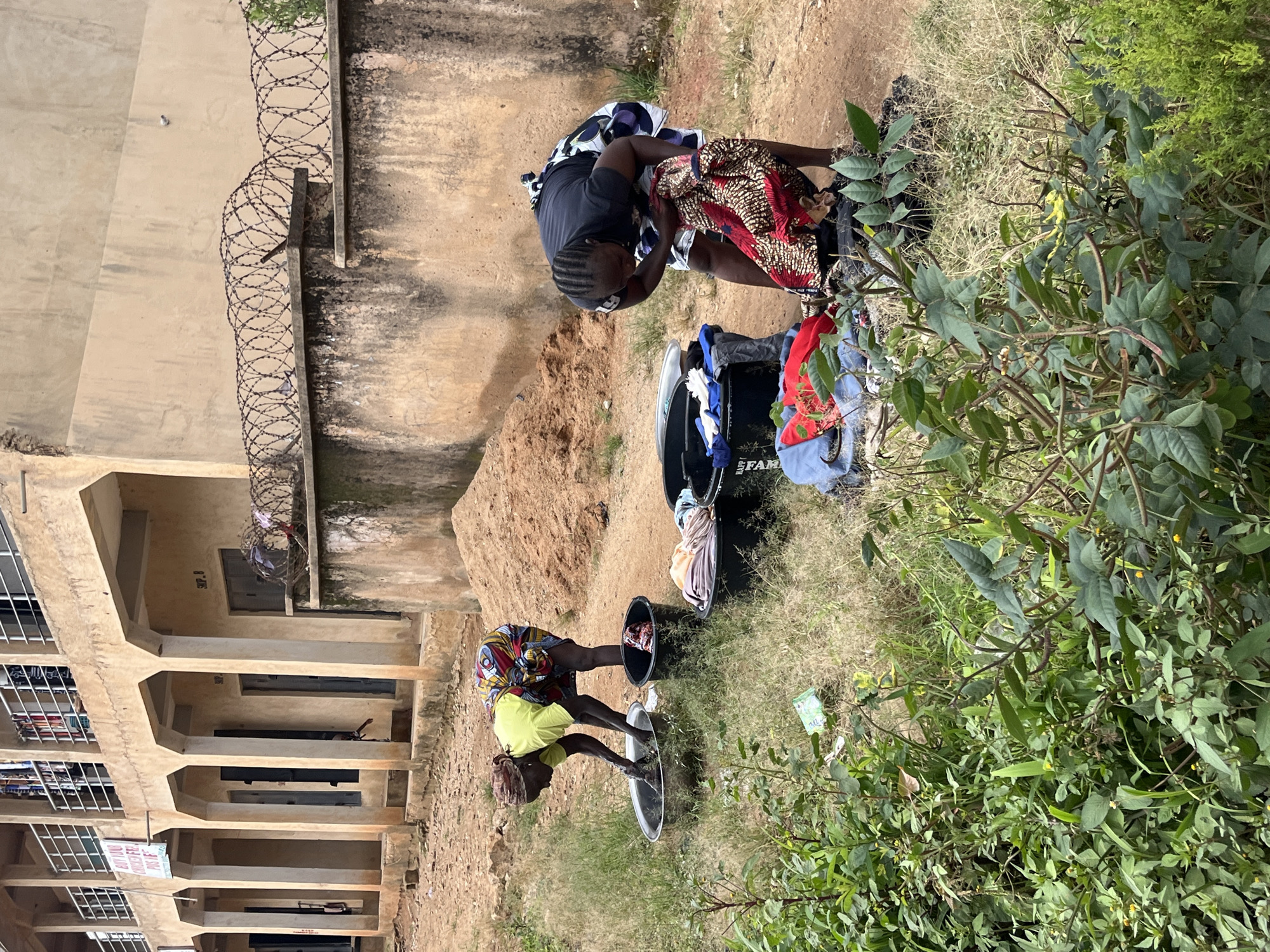 In Bukuru, Nigeria, two women are washing clothes outdoors near a concrete building. The woman on the left, wearing a yellow shirt and a patterned wrap skirt, is bending over a metal basin, her hands in the water. Beside her is a black bucket, also containing clothing. The woman on the right, in a dark shirt and a patterned wrap skirt, is reaching into a black tub filled with clothes. Laundry is spread out on the ground, with various colored fabrics visible. Behind them is a tan building with arched walkways and barred windows, and a wall topped with barbed wire. The ground is a mix of dirt and dry grass, with some green foliage in the foreground.