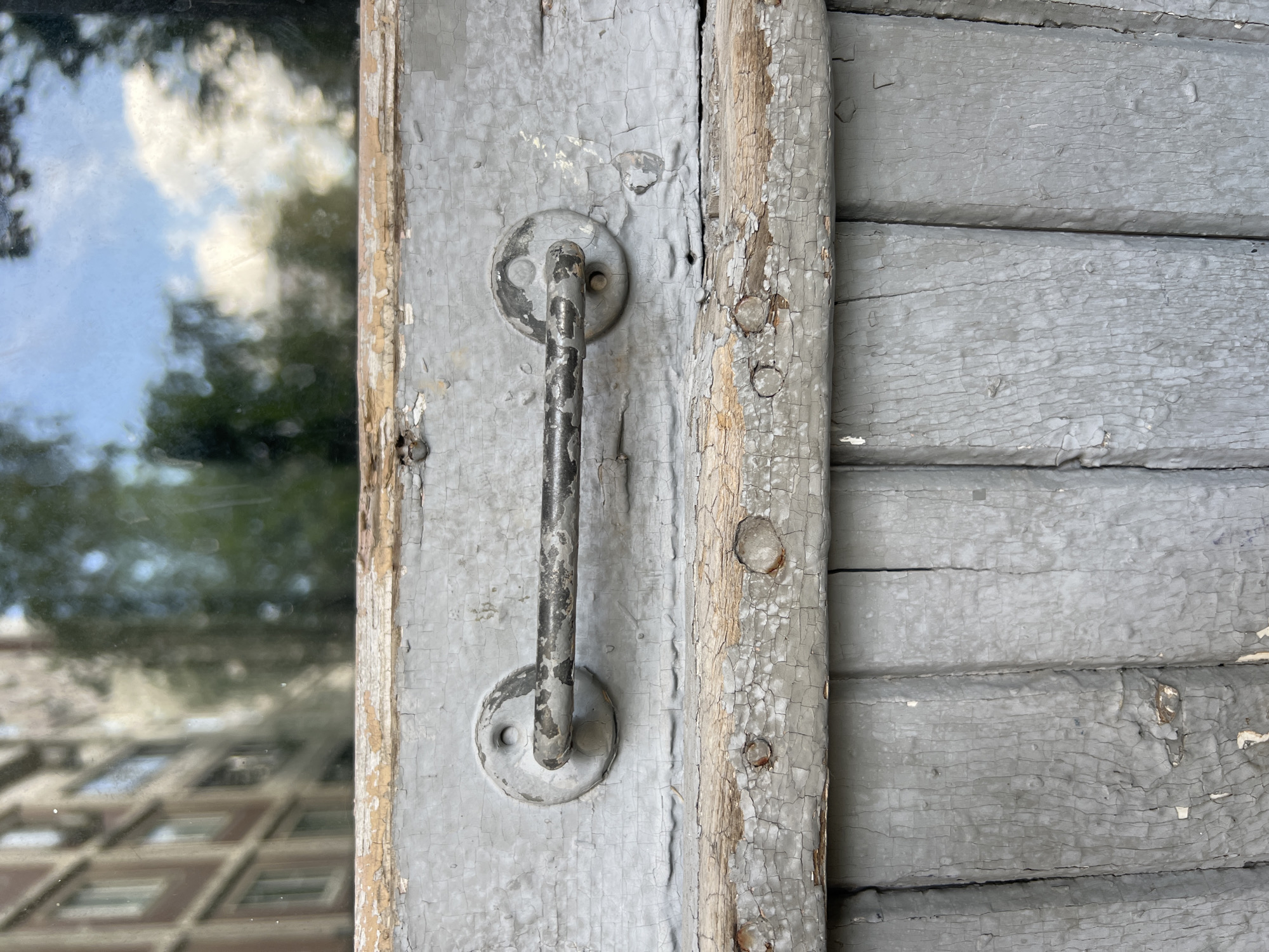 The image shows a close-up of an old, weathered gray wooden door with a metal handle. The paint is chipped and cracked, revealing the wood underneath. In the reflection of the window above the door, a blurred building and trees can be seen, suggesting an outdoor urban environment. The image is taken in Moscow, Russia.