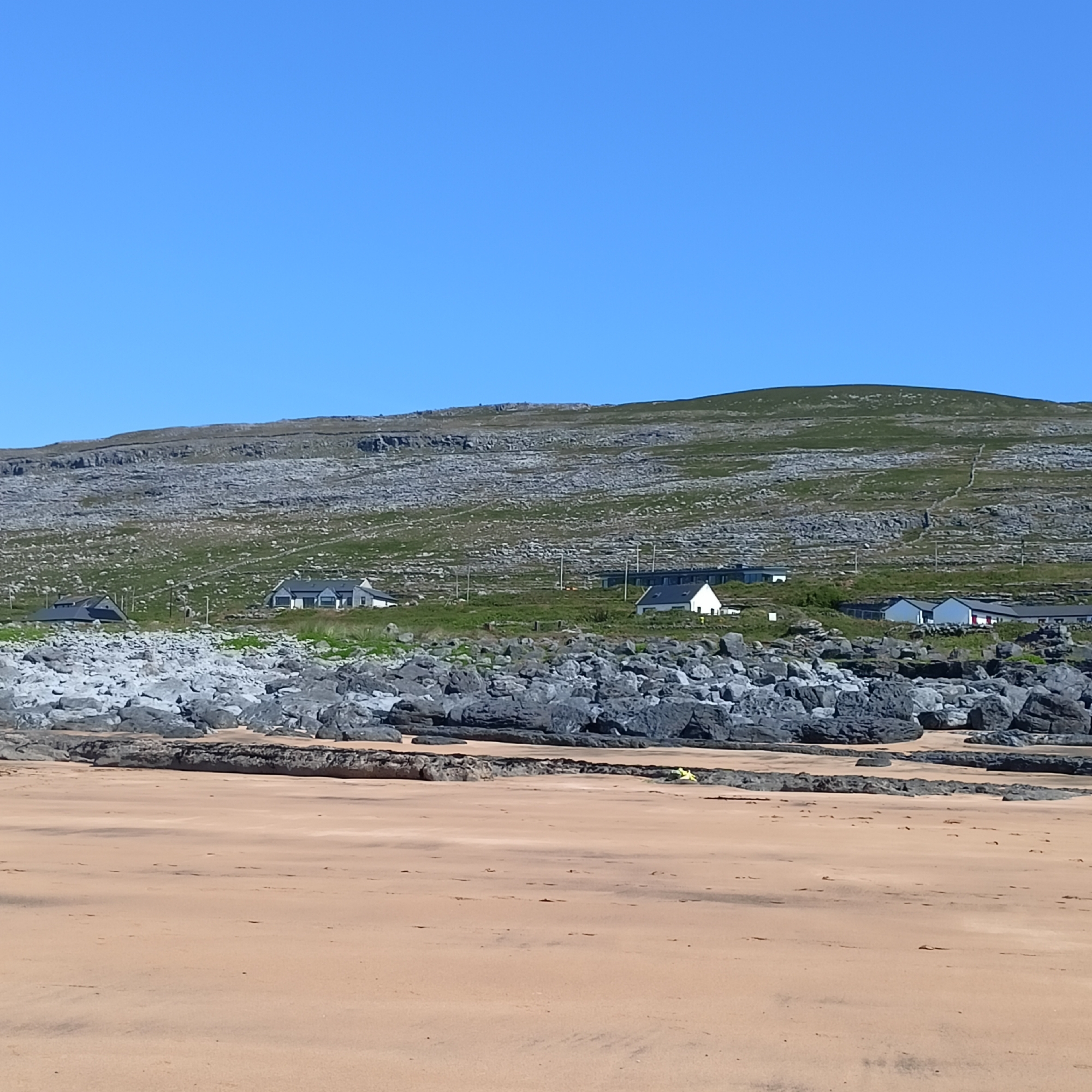 A wide, sandy beach with light brown sand stretches across the foreground. Beyond the beach are large, dark gray rocks and boulders. Further back, the scene transitions to a grassy, rocky hillside dotted with several white houses with dark roofs. The hillside slopes upwards towards a ridge that forms the top of the frame. The sky above is a clear, bright blue. The overall scene depicts a coastal landscape in Fanore, Ireland, under a sunny sky.