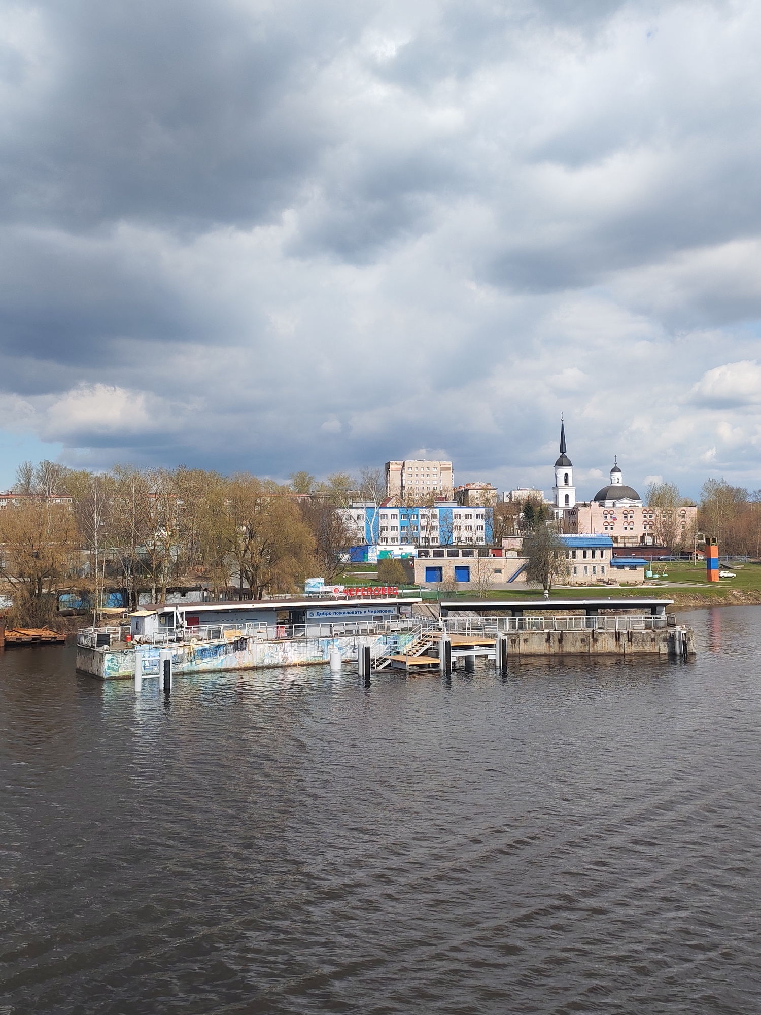 The image shows a waterfront view in Cherepovets, Russia. In the foreground is dark water with visible ripples. A structure, possibly a pier or docking area, extends into the water; it features a flat platform with white supports and colored graffiti-like designs on the side. A staircase leading up to the platform is visible.
In the background, there's a skyline consisting of trees with budding leaves, residential buildings, and what appears to be a church. The church has a tall, slender spire and a rounded dome. The sky above is overcast with dark, heavy clouds.