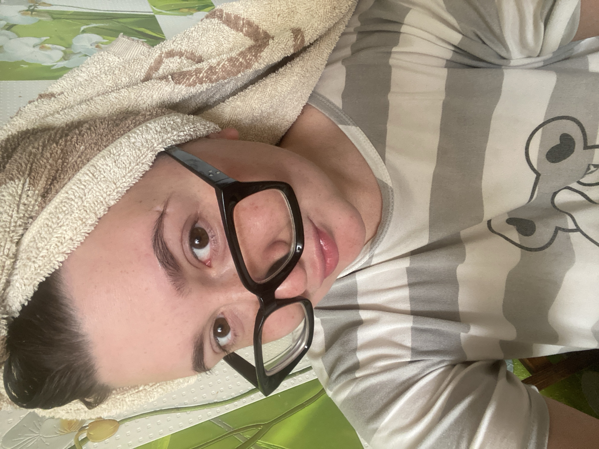 An indoor setting in Chaikovsky, Russia, features a young adult female captured in a close-up, chest-up selfie. Her dark, damp hair is pulled back and wrapped in a light beige terry cloth towel with brown patterned accents, consistent with having recently washed it. She has fair skin with visible natural texture, prominent dark eyebrows, and brown eyes. She wears thick-framed black eyeglasses, with the frames partially obscuring her eyes. Her lips are full and slightly parted, forming a subtle half-smile or pout as she gazes directly at the camera. She is dressed in a horizontally striped T-shirt with alternating light grey and white bands. A graphic depicting a simplified cartoon character, resembling a bear with two heart shapes on its head, is visible on the lower right portion of the shirt. The background consists of a wall covered in light green wallpaper adorned with a white floral pattern, primarily depicting orchids.