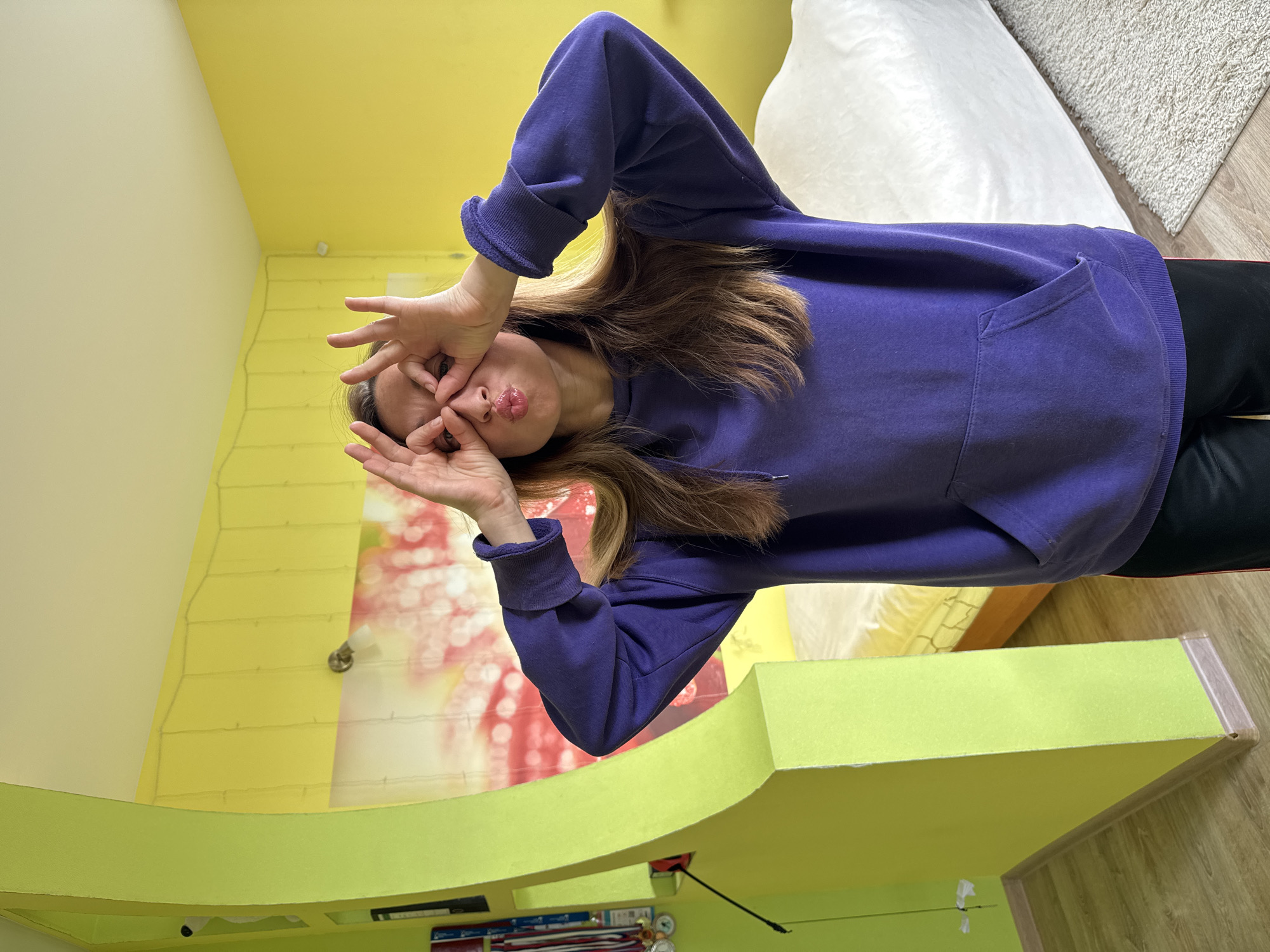 A person with light brown hair and wearing a purple hooded sweatshirt and black pants is making a face while holding their hands up to their face with their fingers and thumbs forming small circles in front of their eyes. The setting is indoors with yellow walls and a curved, lime green ledge or shelf. A white beanbag chair and a wooden floor with a small rug are visible in the background. The lighting appears to be from a combination of natural and artificial sources. The location is Прикамский, Russia.