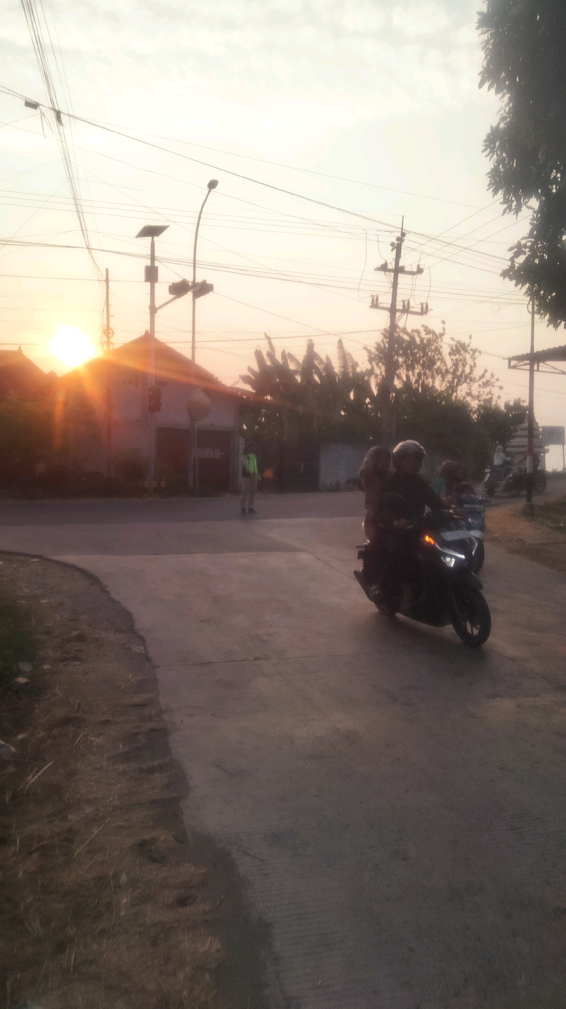 A street scene in Palang, Indonesia, at sunrise. The sun is low on the horizon, casting a warm glow over the sky and illuminating the scene with a golden light. The sky is filled with soft clouds. Several power lines crisscross the sky, supported by utility poles. In the foreground, a paved road curves to the left. On the right side of the road, a dark-colored motorcycle with two riders, both wearing helmets, travels away from the viewer. The rider in front appears to be a male, and the passenger behind is likely a female. The motorcycle's headlight is illuminated. To the left of the road, there is a grassy embankment with dry grass and some sparse vegetation. In the middle ground, a building with a sloped roof is visible, possibly a house or a small shop. A traffic light is attached to a utility pole near the building. A person in a light-colored uniform, possibly a traffic officer or security guard, stands on the side of the road holding a small orange object. Behind the building, trees and foliage are silhouetted against the bright sky. Further in the background, more utility poles and power lines are visible. The overall atmosphere is peaceful and evokes the early morning hours in a suburban or rural setting in Indonesia.