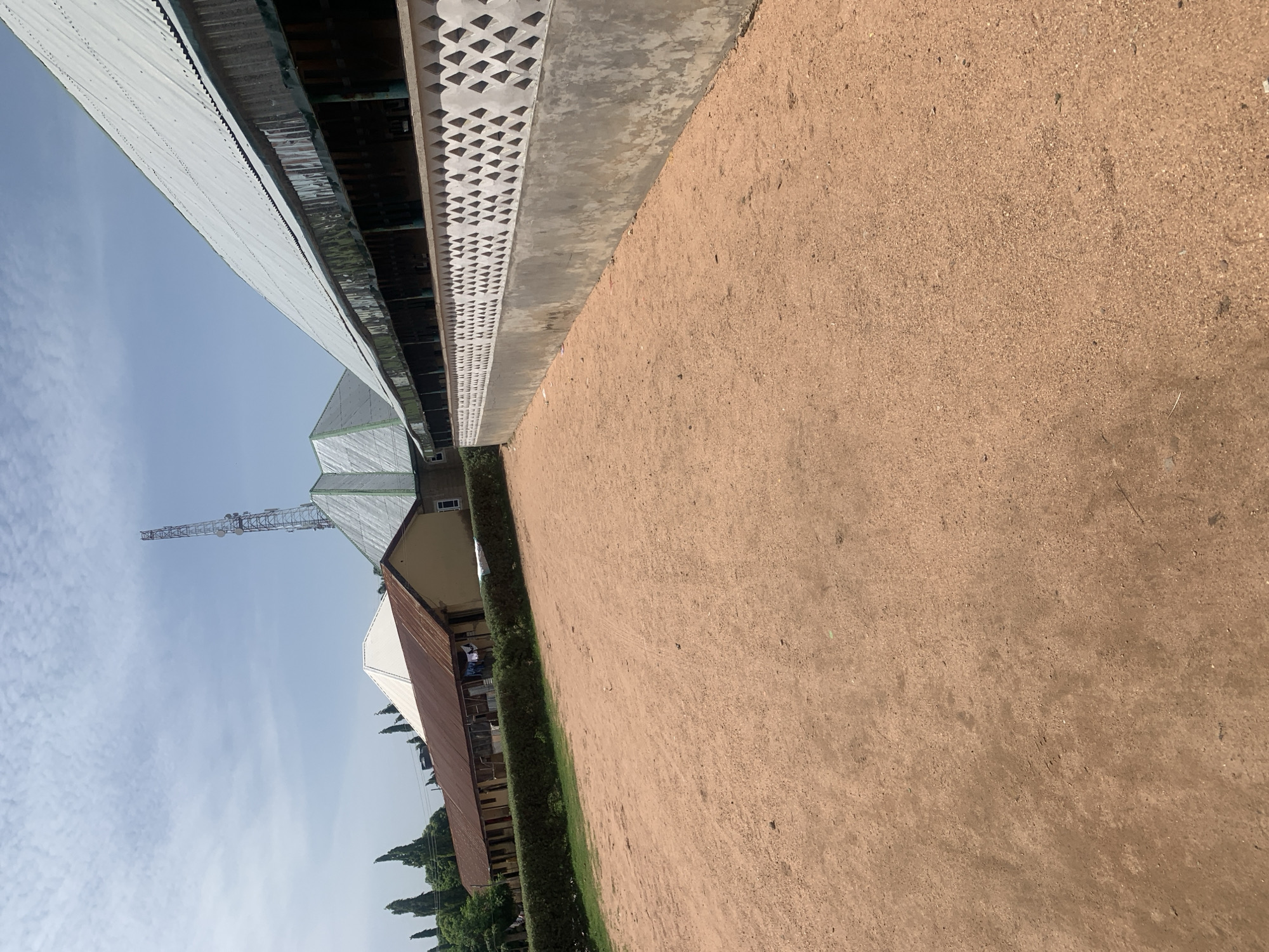 In Jauro Yinu, Nigeria, this image depicts an outdoor scene. The foreground is dominated by a wide expanse of reddish-brown dirt or sand. To the right, a concrete wall with a decorative pattern of diamond-shaped openings runs along the edge of the dirt area. Beyond this wall, a long building with a corrugated metal roof is visible. This building features an open-air, covered walkway along its front. Further in the background, a series of structures with different roof types, including corrugated metal and what appears to be white sheeting, are present. A tall telecommunications tower stands prominently in the background, reaching towards a sky with scattered clouds. Trees, including tall, slender ones, are visible behind the buildings. The overall impression is one of a semi-urban or rural environment, possibly a school or community complex, under a bright, daytime sky. There are no people or animals visible in the image.