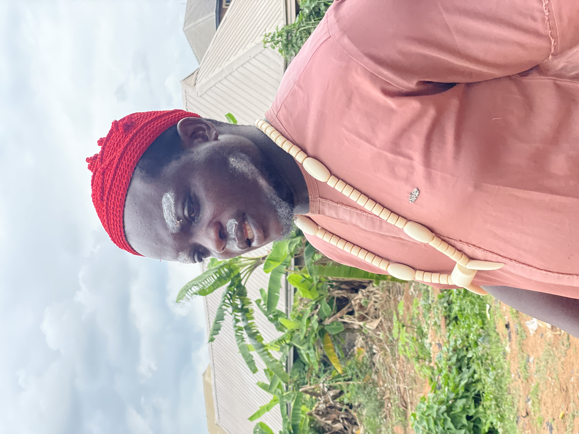 A dark-skinned man is depicted in a vertical, waist-up outdoor portrait, looking slightly upward and to his left. He is wearing a vibrant red, knitted cap that covers his head. White markings, resembling chalk or paint, are visible on his forehead and below his right eye. He wears a light reddish-brown long-sleeved shirt, which features a small, intricate metal emblem on the left side of his chest. Around his neck hangs a prominent necklace made of numerous elongated, light-colored beads, with a larger, oval central bead and two pointed, tooth-like embellishments at the front. His expression is neutral, with his mouth slightly open.
The background shows a mix of natural and man-made elements. Lush green foliage, including broad banana-like leaves, is visible, alongside areas of dry, reddish-brown soil. In the upper background, structures with light-colored corrugated metal roofs suggest residential or commercial buildings. The sky is largely overcast with soft, grey clouds, indicating a cloudy daytime. The attire and markings on the man suggest cultural or traditional significance, fitting the context of Onitsha, Nigeria. No visible text is present in the image.