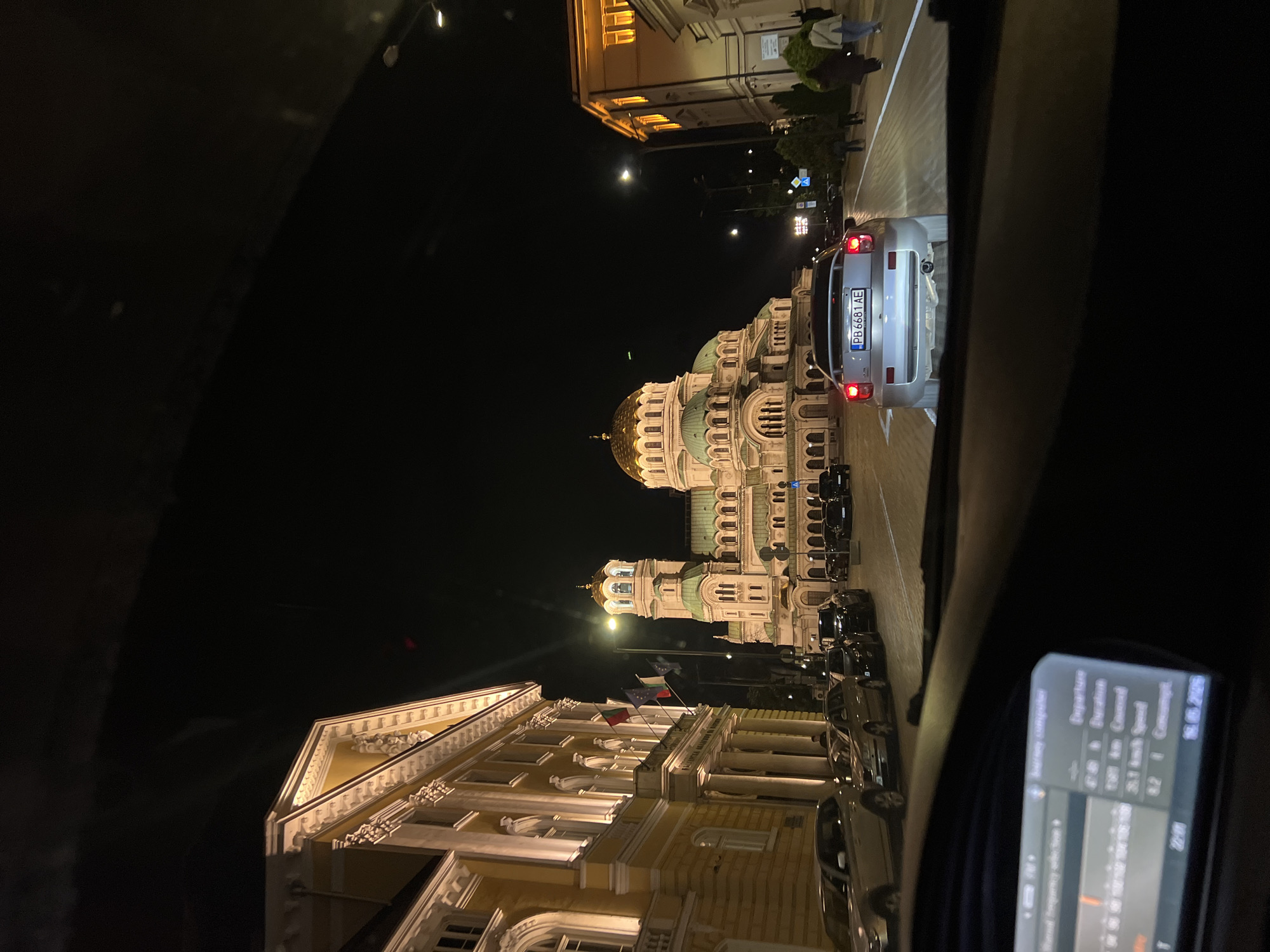 Captured from within a vehicle at night, the image presents a street scene in Sofia, Bulgaria. The Alexander Nevsky Cathedral, an elaborate Eastern Orthodox cathedral with a gilded dome, is prominently featured. Adjacent to the cathedral is an ornate, yellow-toned building flying the Bulgarian flag, and possibly the flag of the European Union, and is likely the Bulgarian Parliament. The camera is focused on a vehicle directly in front of the photographer's car, illuminated by its tail lights and featuring license plate number "PB 6681 AE". On the right side of the frame, a dashboard screen displays a navigation system, showing journey computer information including departure, duration, and average speed. Pedestrians can be seen walking on the sidewalks. Street lamps illuminate the architecture.