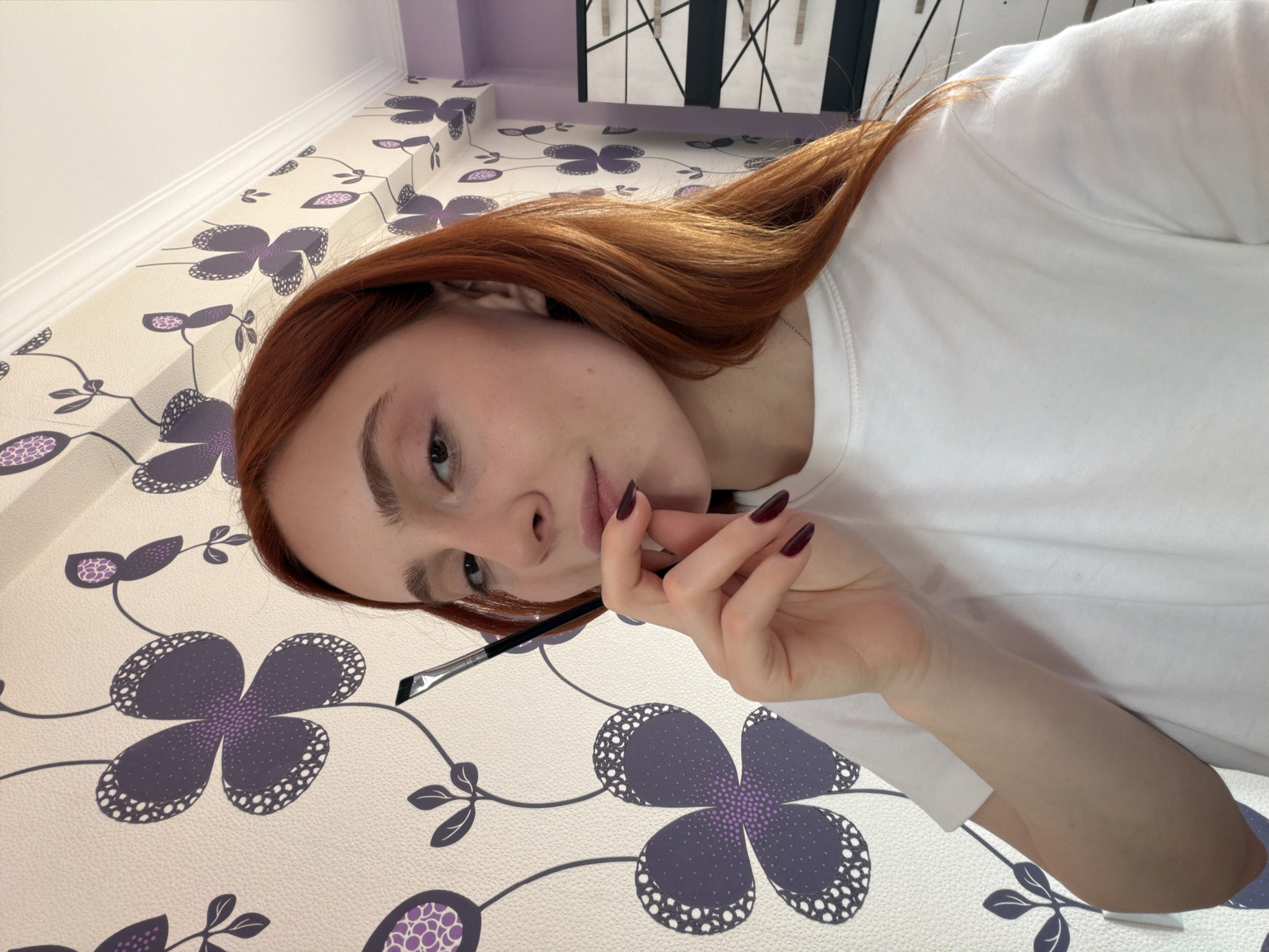 A young, red-haired woman wearing a white t-shirt is holding a slanted-tip makeup brush near her face, against a background of purple floral wallpaper. The setting includes a room with visible architectural elements. The location cannot be determined from the image.