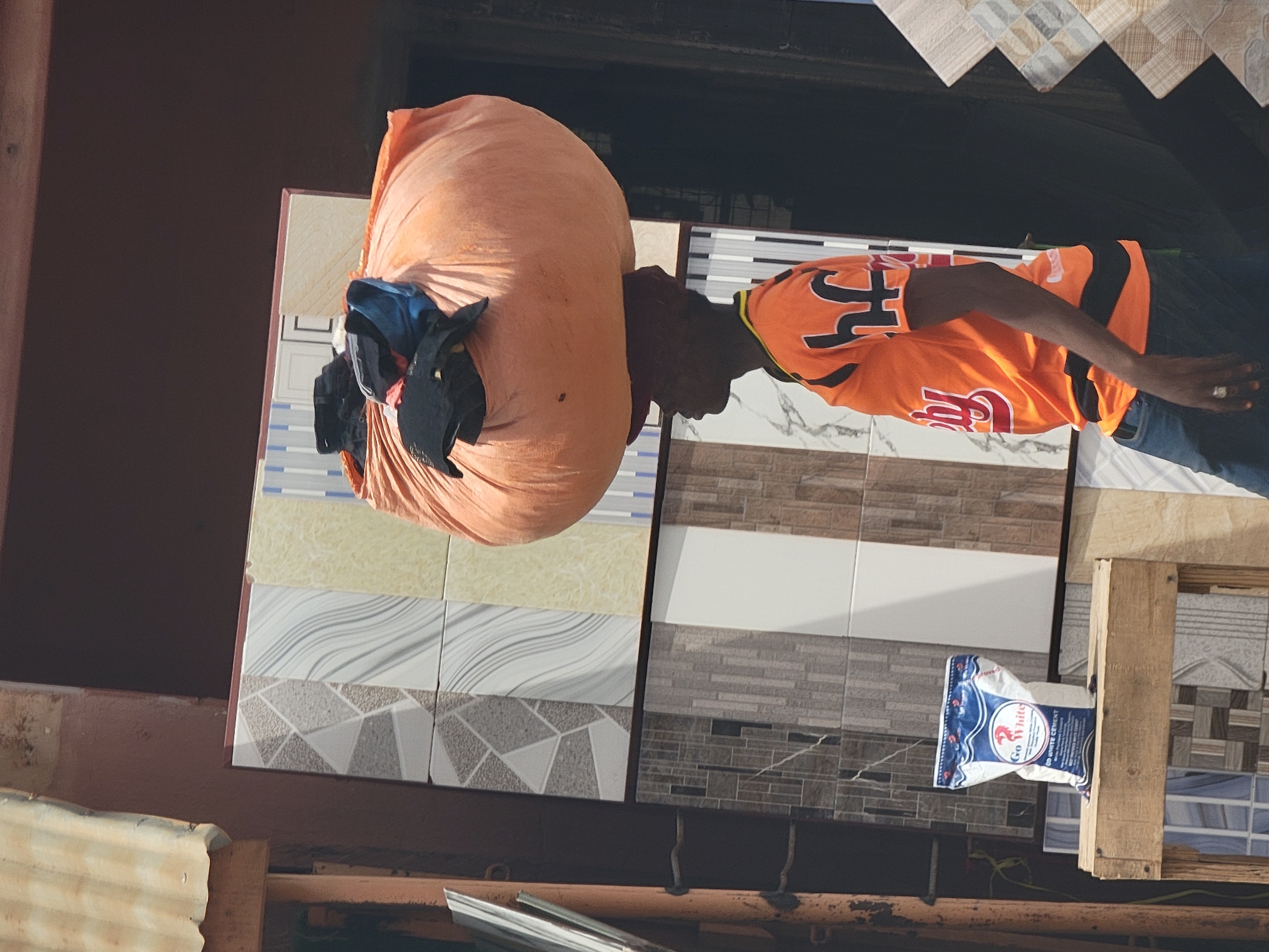 A man in an orange shirt carries a large orange sack on his back in Maiburiji, Nigeria. The sack contains several items of clothing. He stands next to a display of various ceramic tiles, with a bag of "Go White Cement" visible nearby. The surrounding environment includes metal structures and what appears to be parts of buildings.